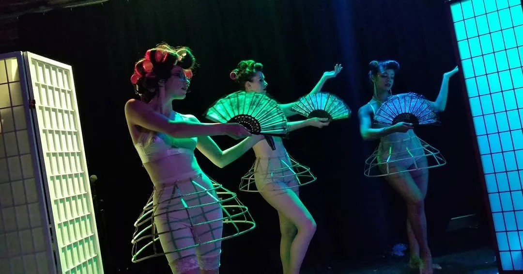 Three women in futuristic, cage-like skirts and neon hair rollers holding decorative fans, performing on a stage with colorful lighting.