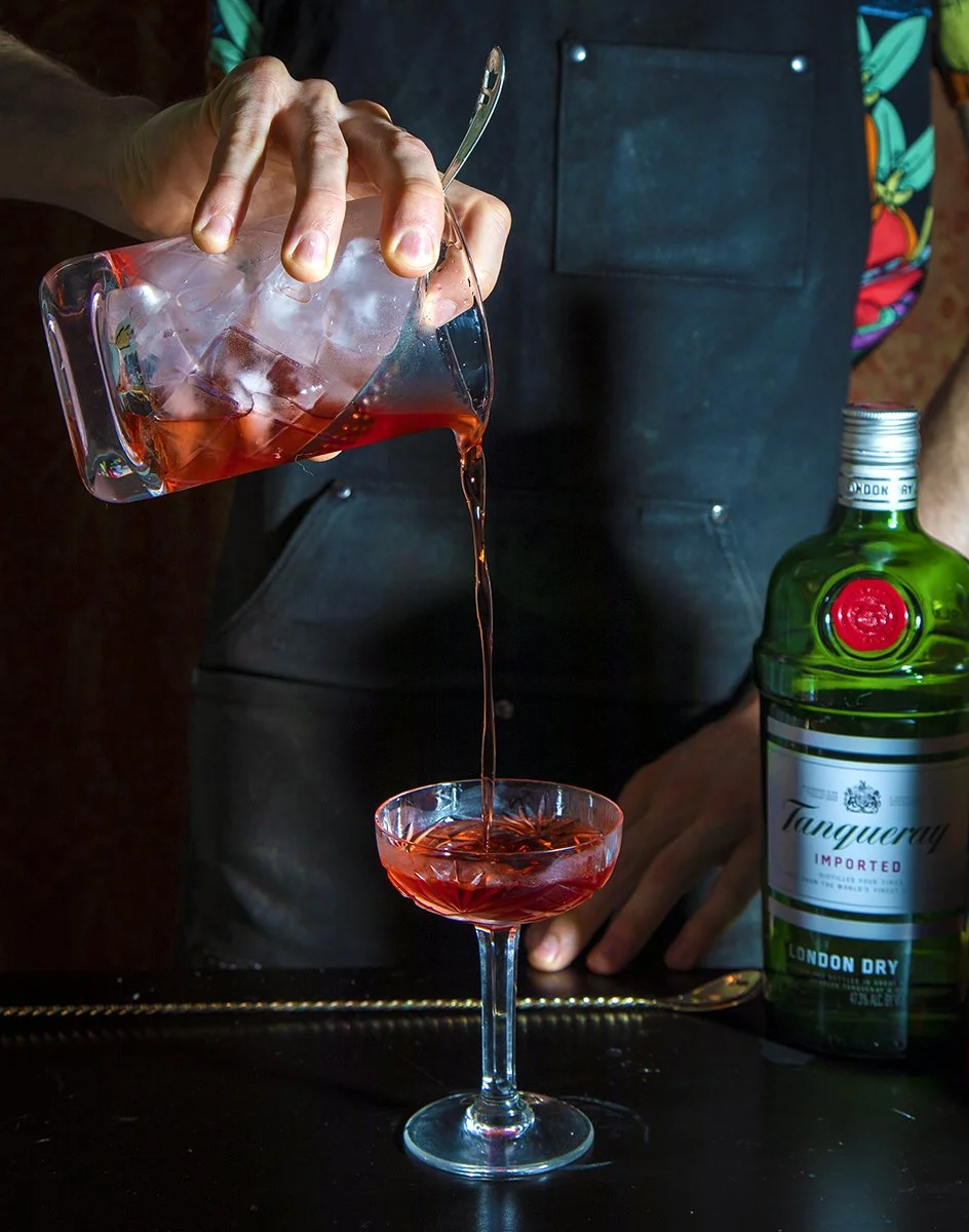 Person pouring red liquor from a glass pitcher into a cocktail glass, with a green bottle of Tanqueray gin on the side.