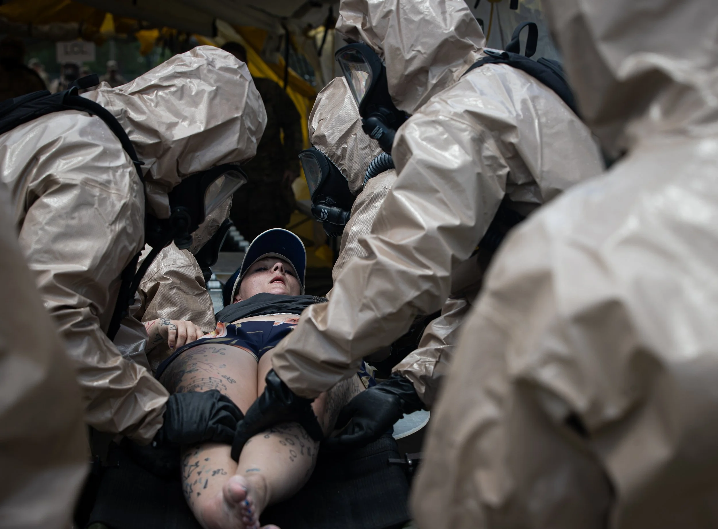 Close-up of a person lying on a stretcher with tattoos on their legs, surrounded by individuals in beige hazmat suits and face masks, possibly paramedics or rescue workers.