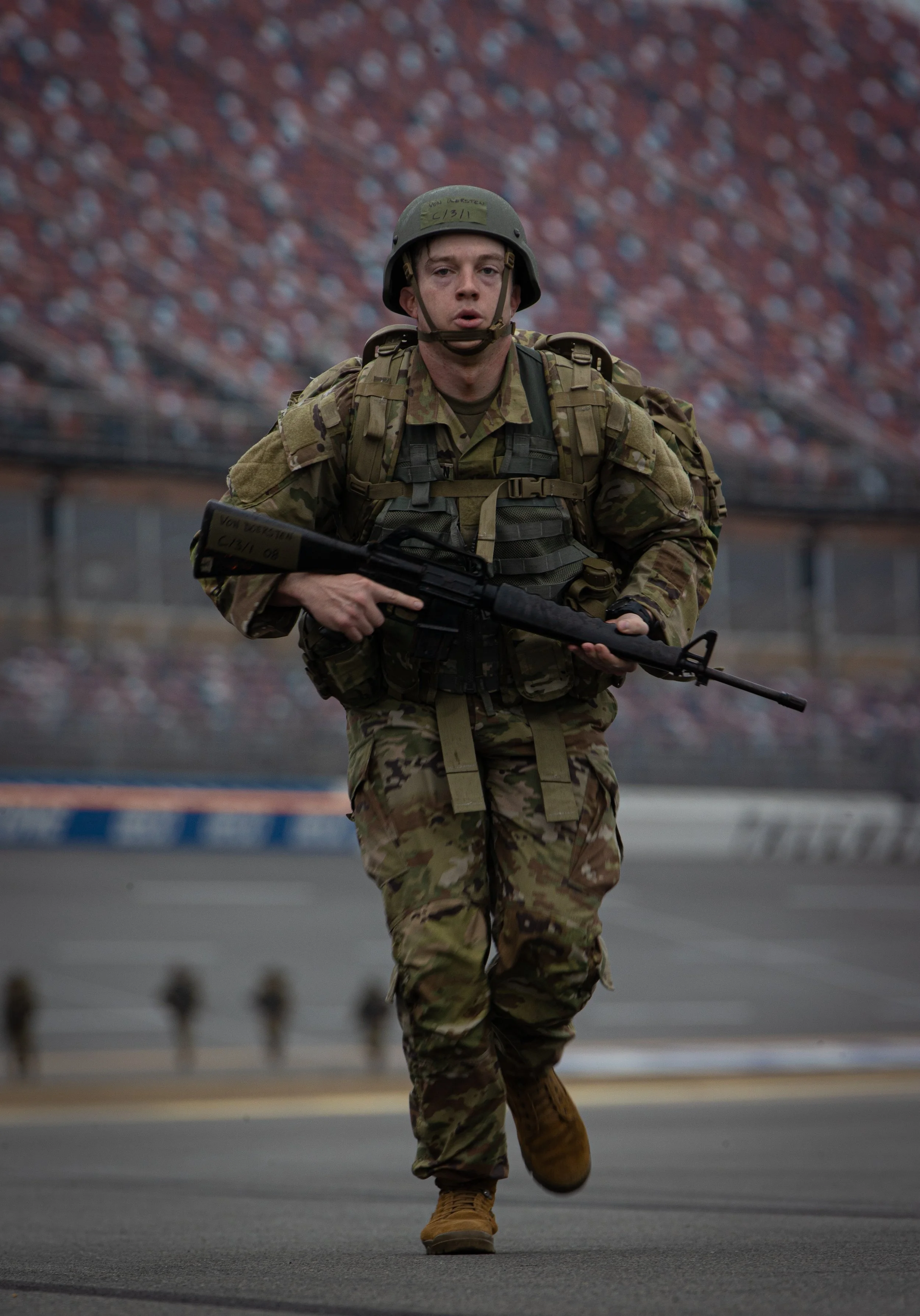A soldier in camouflage uniform and helmet running towards the camera on a paved surface, with an empty stadium in the background.