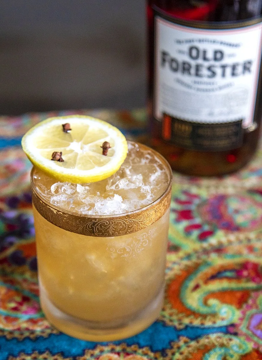 A glass of whiskey sour garnished with a lemon slice and cloves, sitting on a colorful fabric surface with a bottle of Old Forester bourbon in the background.
