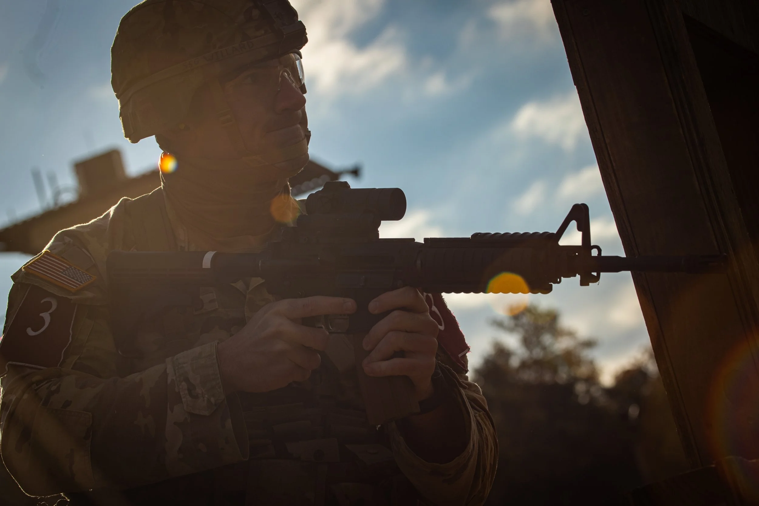 A soldier in camouflage uniform and helmet aiming a rifle through a window or door with a clear sky background.