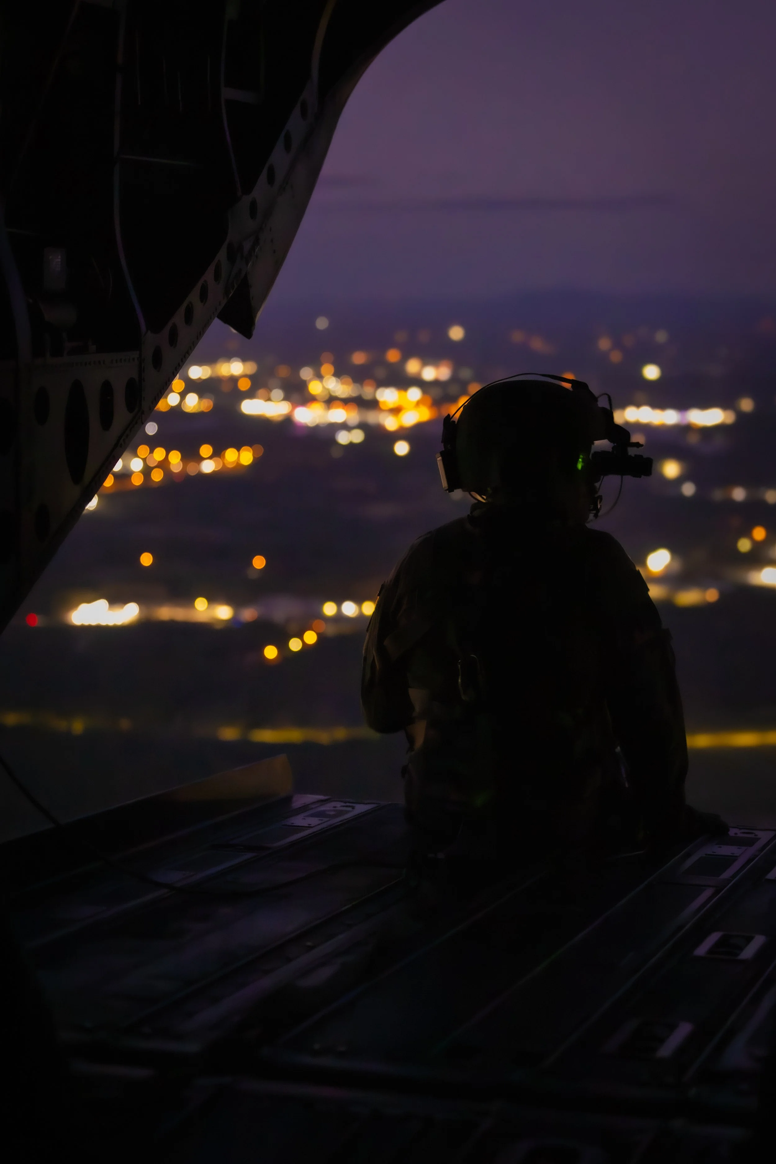 A person wearing a headset inside an aircraft, looking out at the night city lights below, with part of the aircraft's interior visible in the foreground.