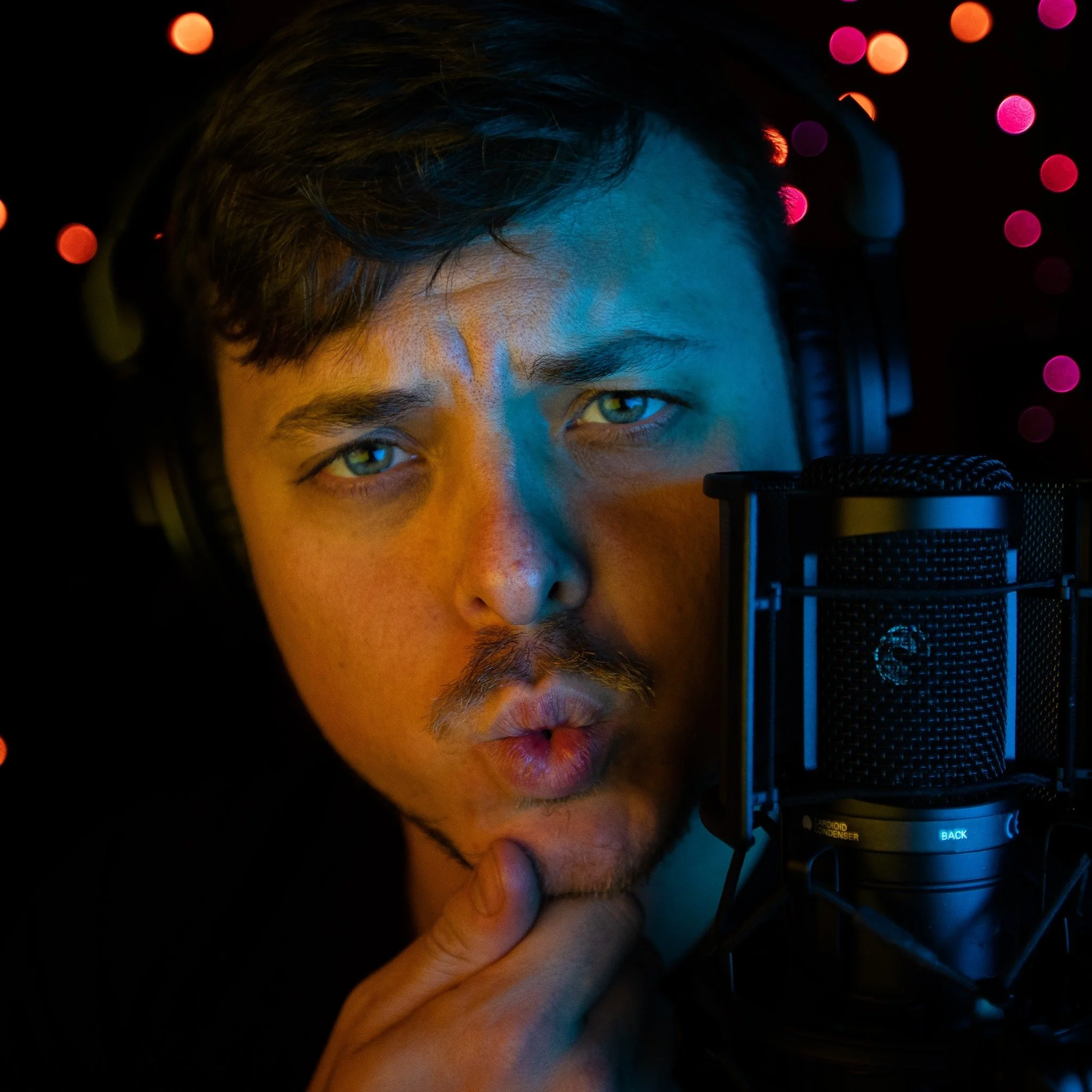 Close-up of a person's face with short hair looking into a professional microphone with headphones, lit by colorful lighting with pink and orange bokeh in the background.