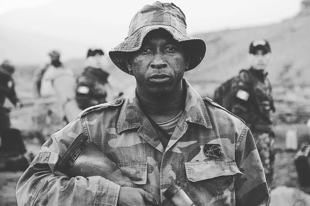 Black and white photo of a soldier in camouflage uniform and hat, holding a weapon, with other soldiers in the background.