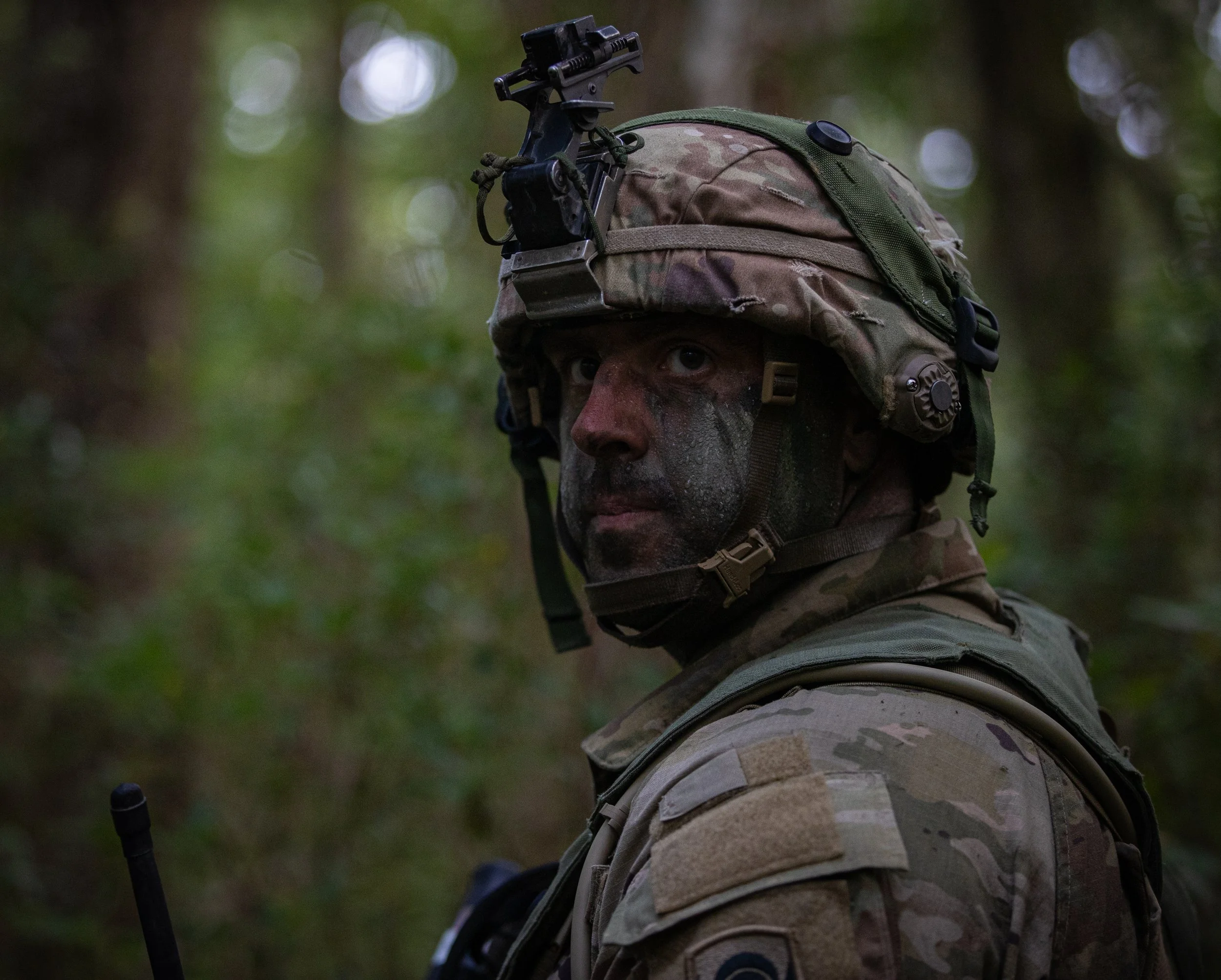 Close-up of a soldier wearing camouflage uniform and helmet with night vision goggles attached, in a forest setting.