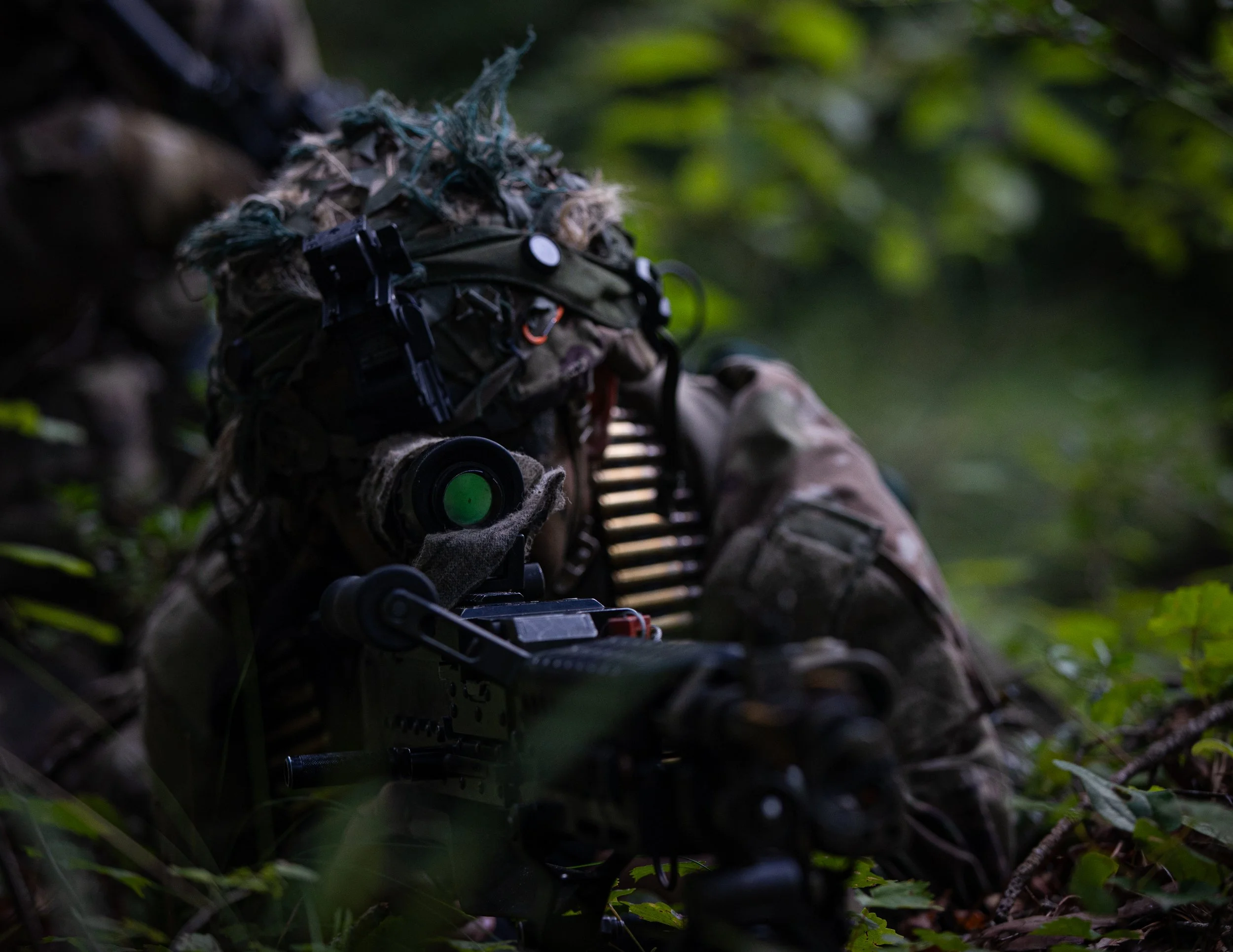A soldier lying on the ground in a forested area aiming a rifle, wearing camouflage gear and a ghillie suit with a scope and tactical equipment.