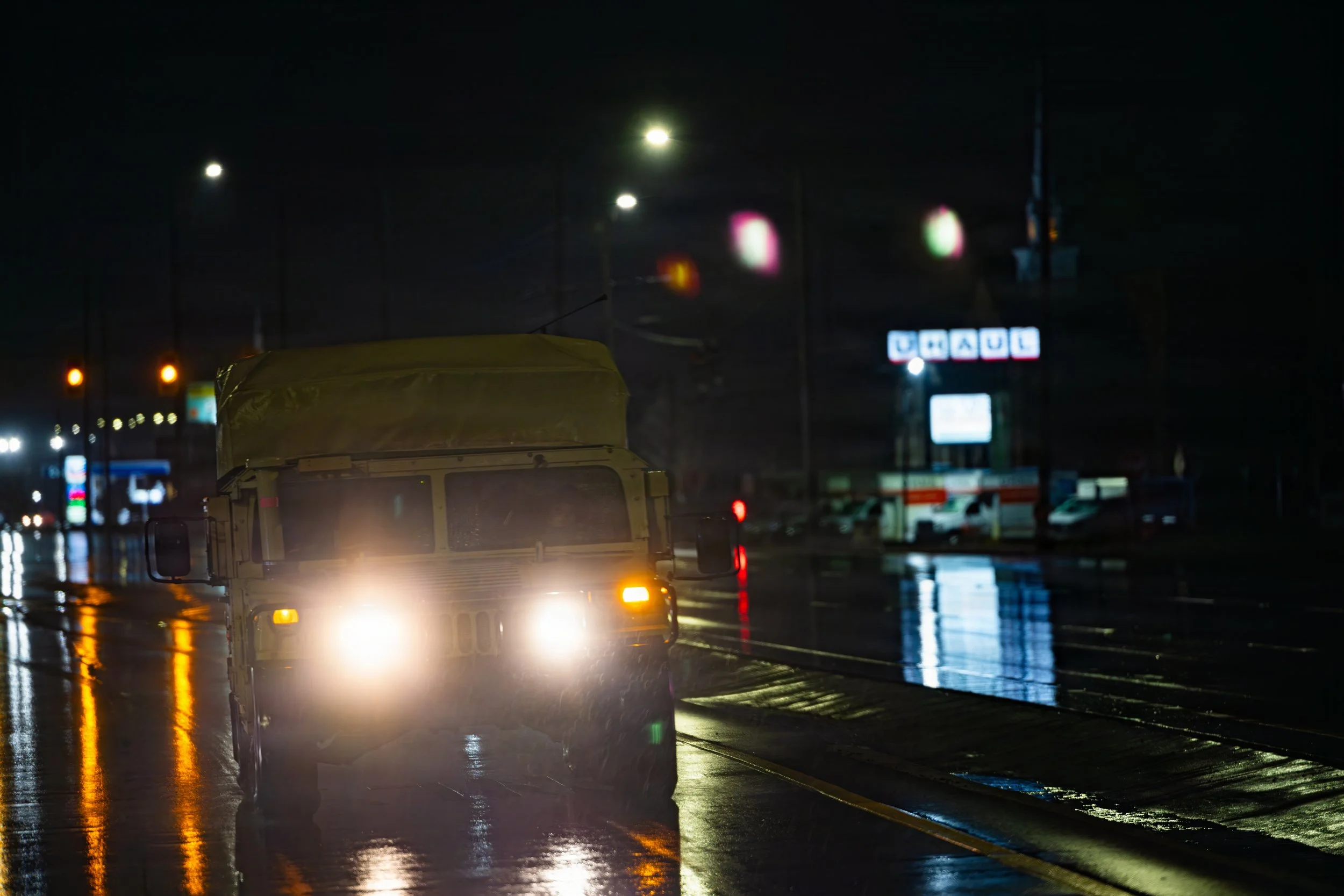 A vehicle driving on a wet road at night with headlights on and streetlights illuminating the scene, reflecting on the rain-soaked pavement.