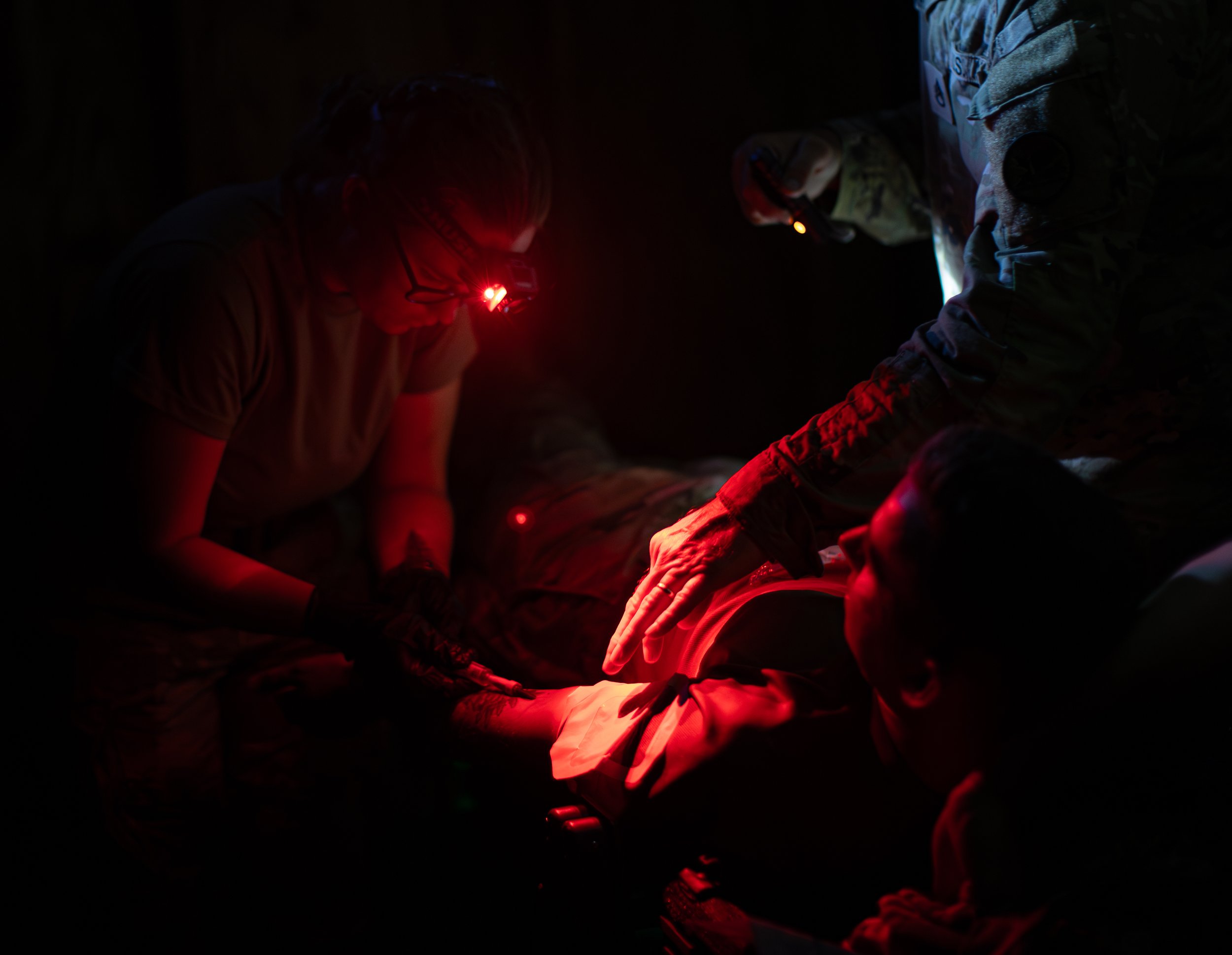 Dark scene with two individuals illuminated by red light, focusing on a person wearing glasses with a headlamp, possibly engaged in a nighttime activity or rescue operation.