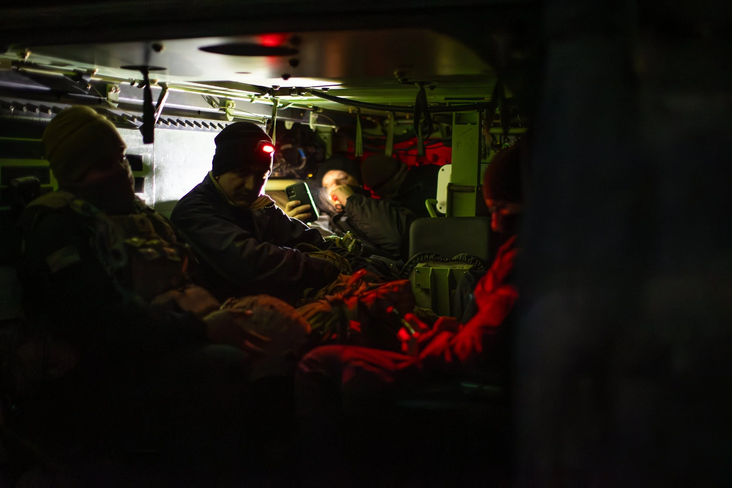 Military personnel inside a dimly lit vehicle, illuminated by a red light, with some individuals using electronic devices.