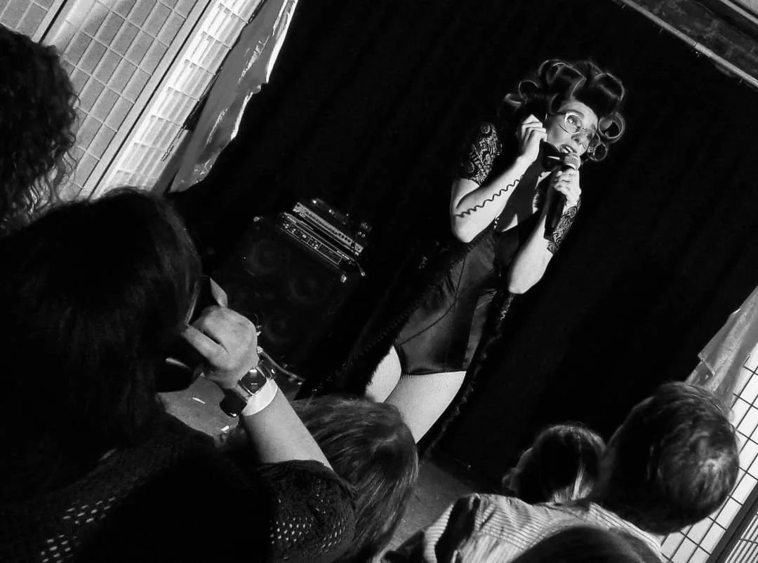 A performer on stage, dressed in a revealing costume with curlers in her hair, singing into a microphone in front of an audience in a dimly lit room.