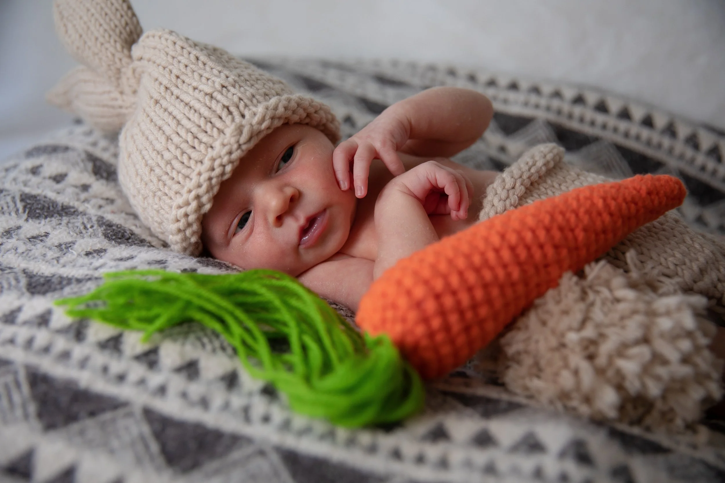 Close-up of a newborn baby wearing a beige knitted hat and dressed in a beige knitted outfit, lying on a patterned blanket with a green yarn ball nearby.