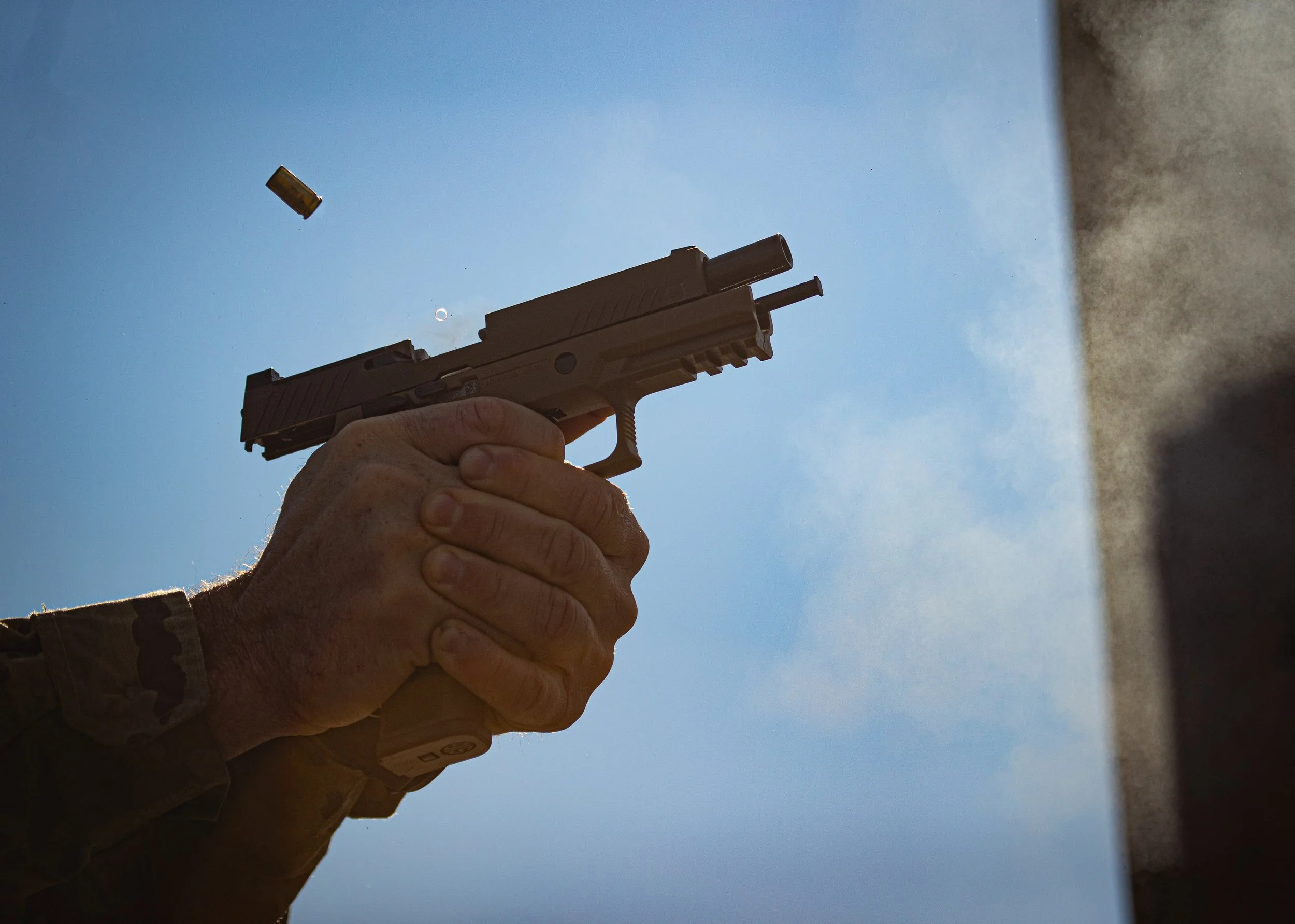 A person with a camo sleeve holding a handgun, firing it at the sky, with shell casings ejecting mid-air.
