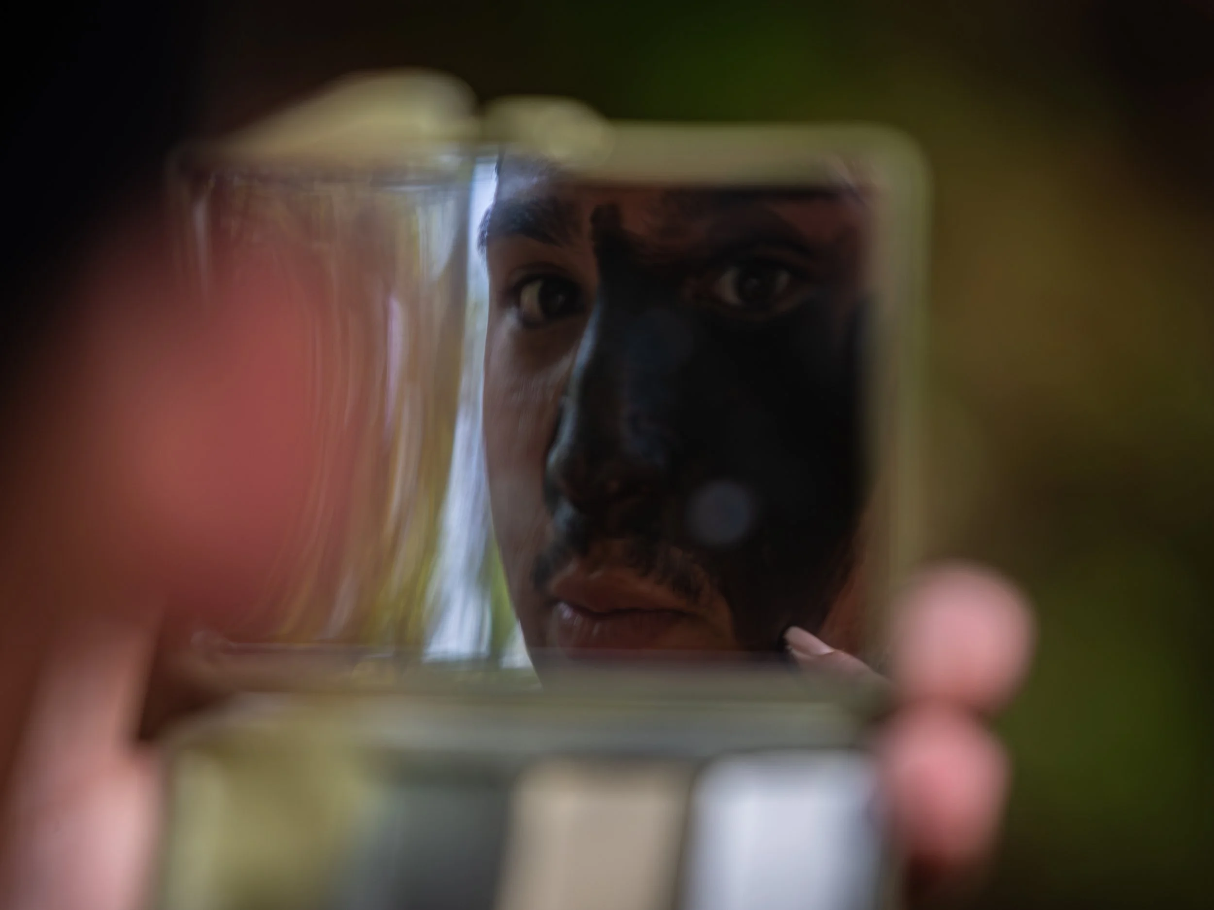 Close-up of a person's face seen through a glass, with a hand partially covering the lens, creating a blurred, intimate perspective.