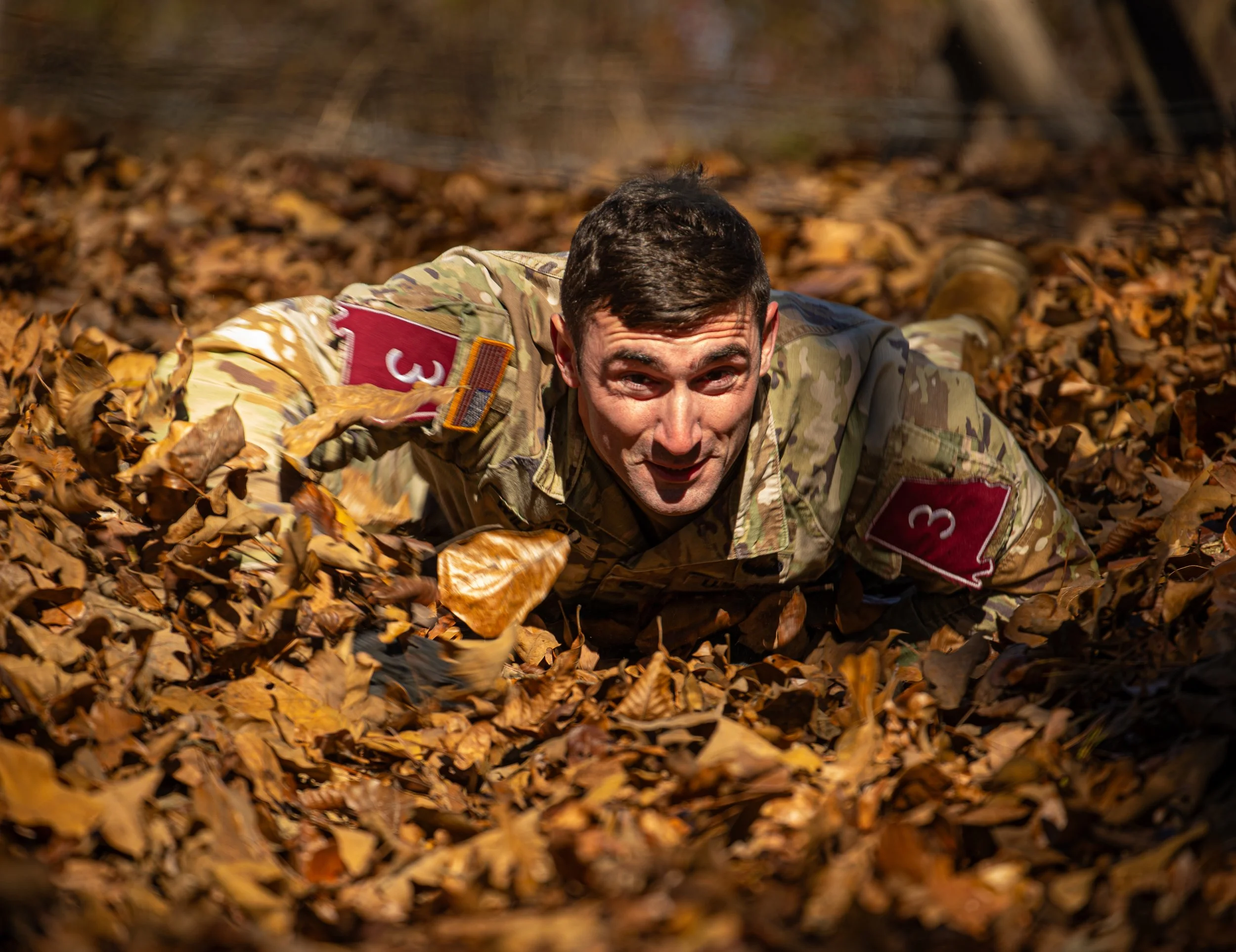 A man in military camouflage clothing crawling through fallen autumn leaves in a forest.