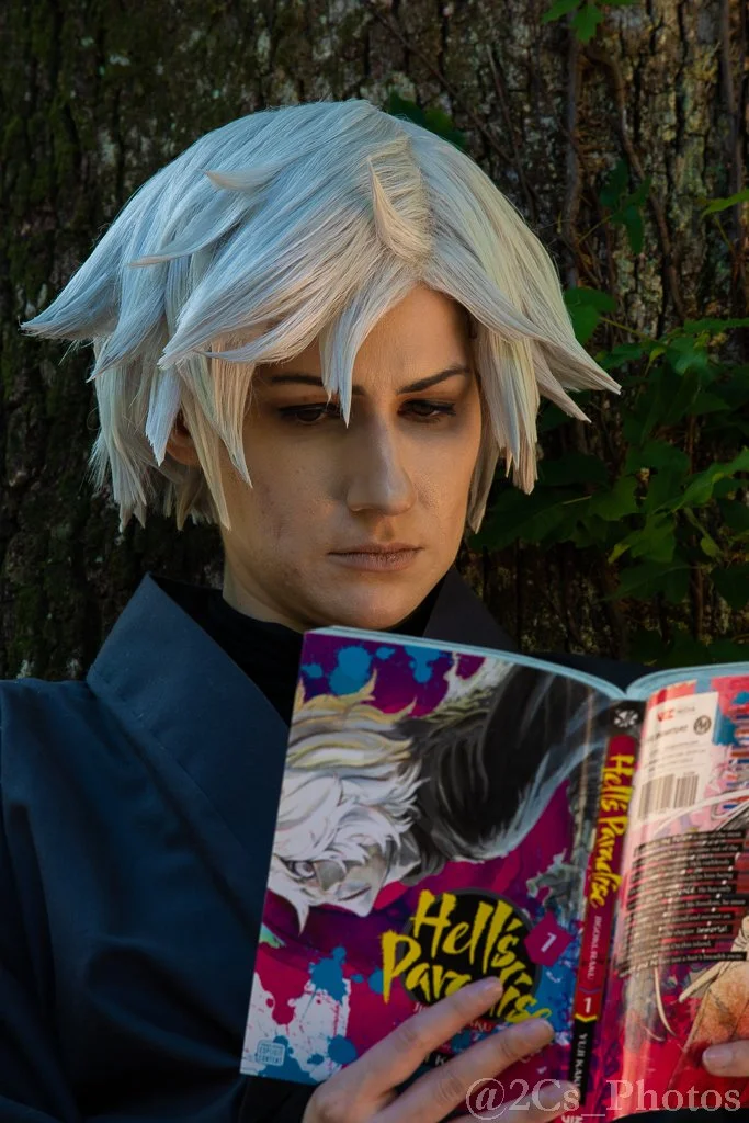 A person with short, spiky, platinum blonde hair styled with several small, upward-pointing horns, wearing a dark jacket, and reading a comic book titled 'Hells Paradise' outdoors with a tree and green foliage in the background.