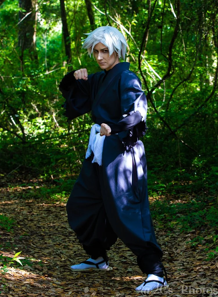 Person dressed in black traditional martial arts attire with white hair wig, standing in a green forest with a serious expression, striking a martial arts pose.