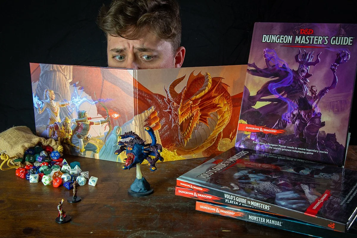 A person with brown hair and blue eyes looks at several D&D game materials on a wooden table, including a Dungeon Master's Guide, a Monster Manual, and a Volo's Guide to Monsters. There are miniatures of a blue creature and a female figure, along wit