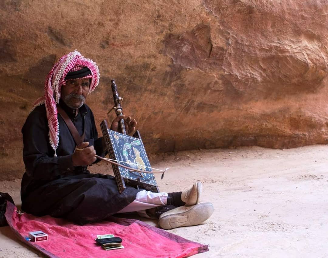 An elderly man wearing traditional Middle Eastern attire, including a red and white checked keffiyeh, sitting on a pink rug in a rocky, cave-like setting. He is playing a musical instrument with a framed artwork in front of him.