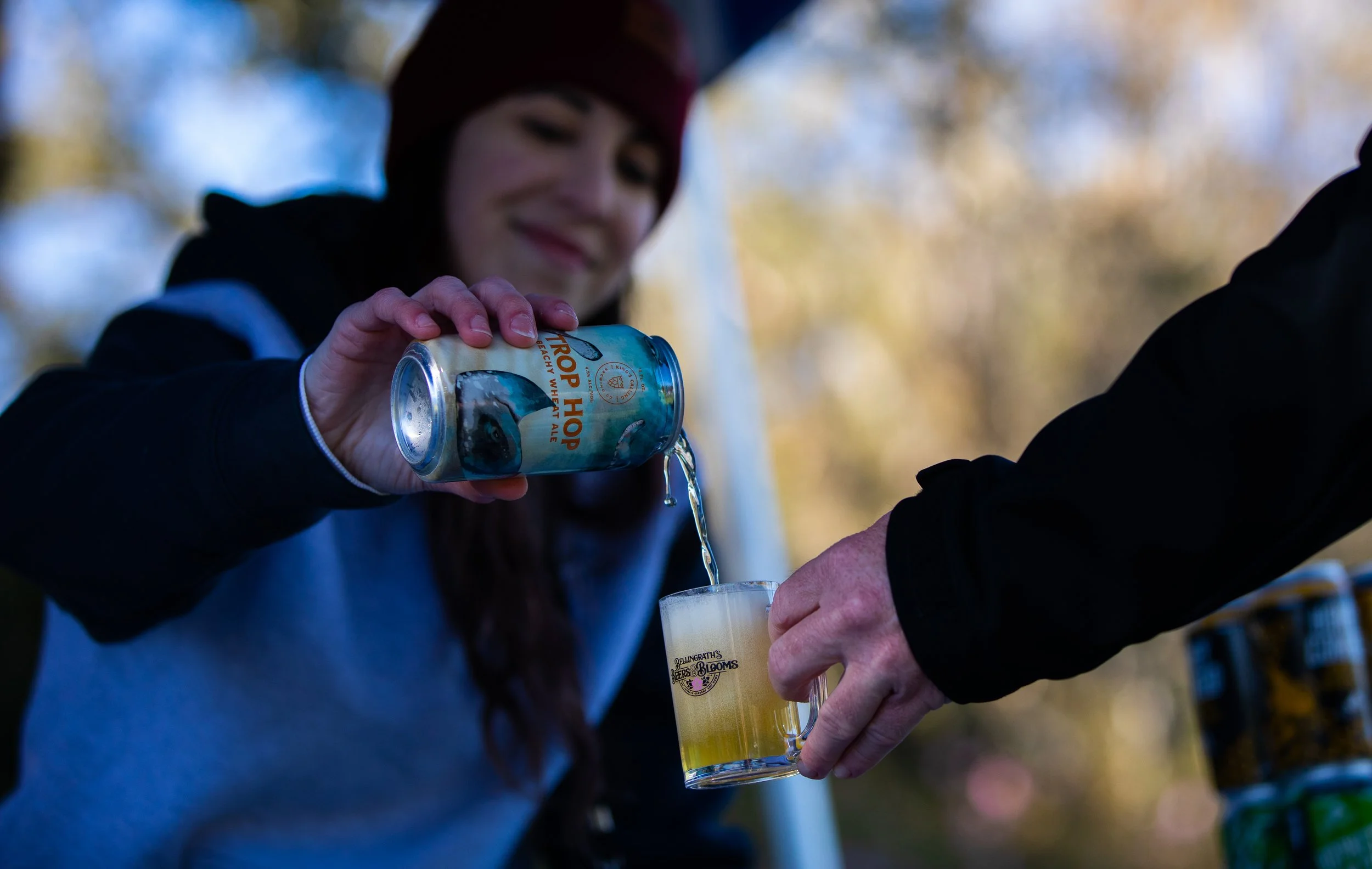 A woman smiling and pouring beer from a can into a glass held by another person outdoors with trees in the background.