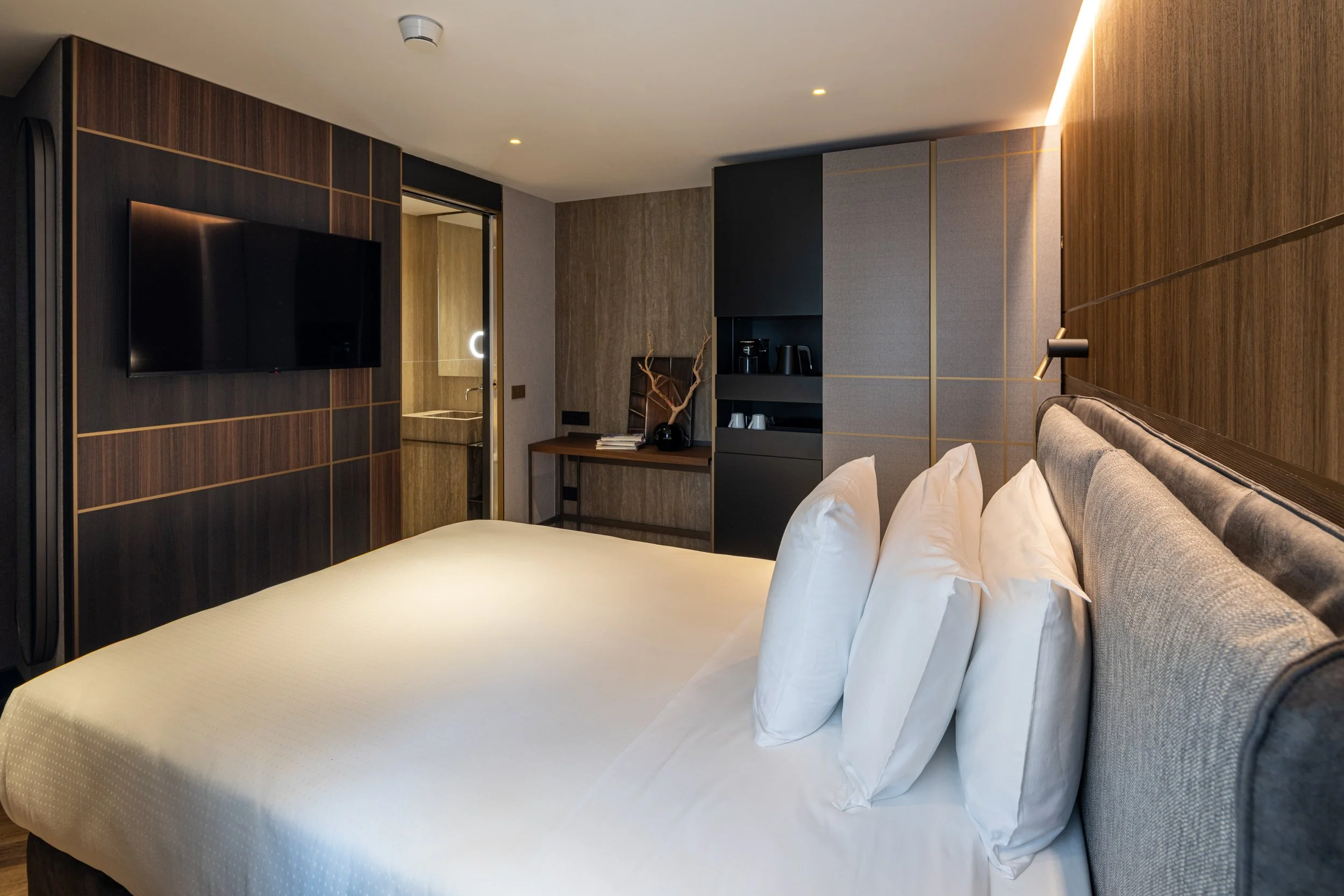 Modern hotel room interior with a bed, gray upholstered headboard, white pillows, wooden and gray wall decor, mounted TV, and a small kitchenette in the background.