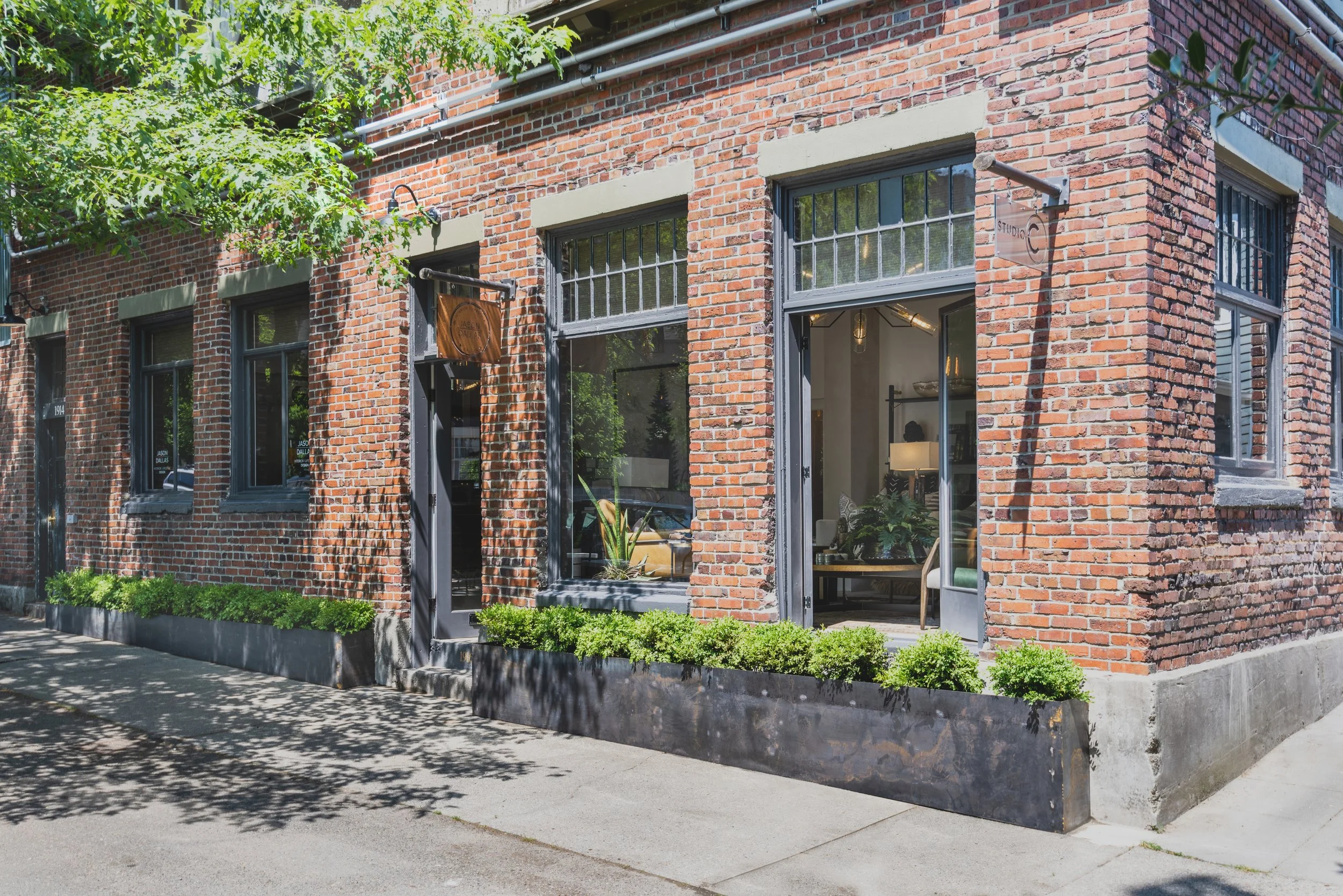 Exterior view of a red brick building with large windows and black metal frames, partially open, showing a warmly lit interior with furniture and plants. Green shrubbery lines the sidewalk in front, and a tree casts shadows on the ground. Decorative signs are mounted on the building exterior.
