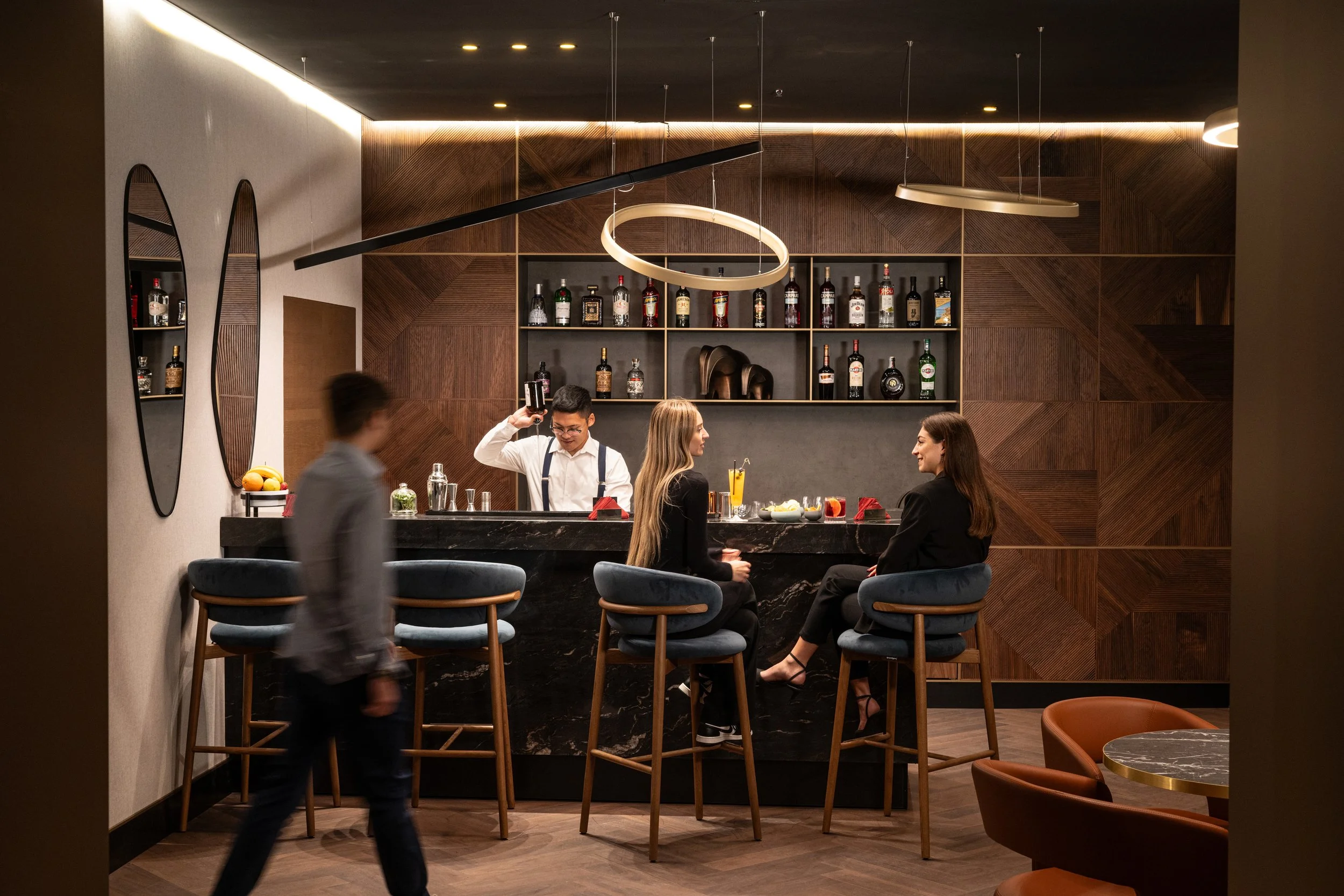 A bar scene with a man pouring a drink behind the counter, two women sitting on bar stools talking, and a blurred man walking past. The background features dark wood paneling with bottles of alcohol displayed on shelves.