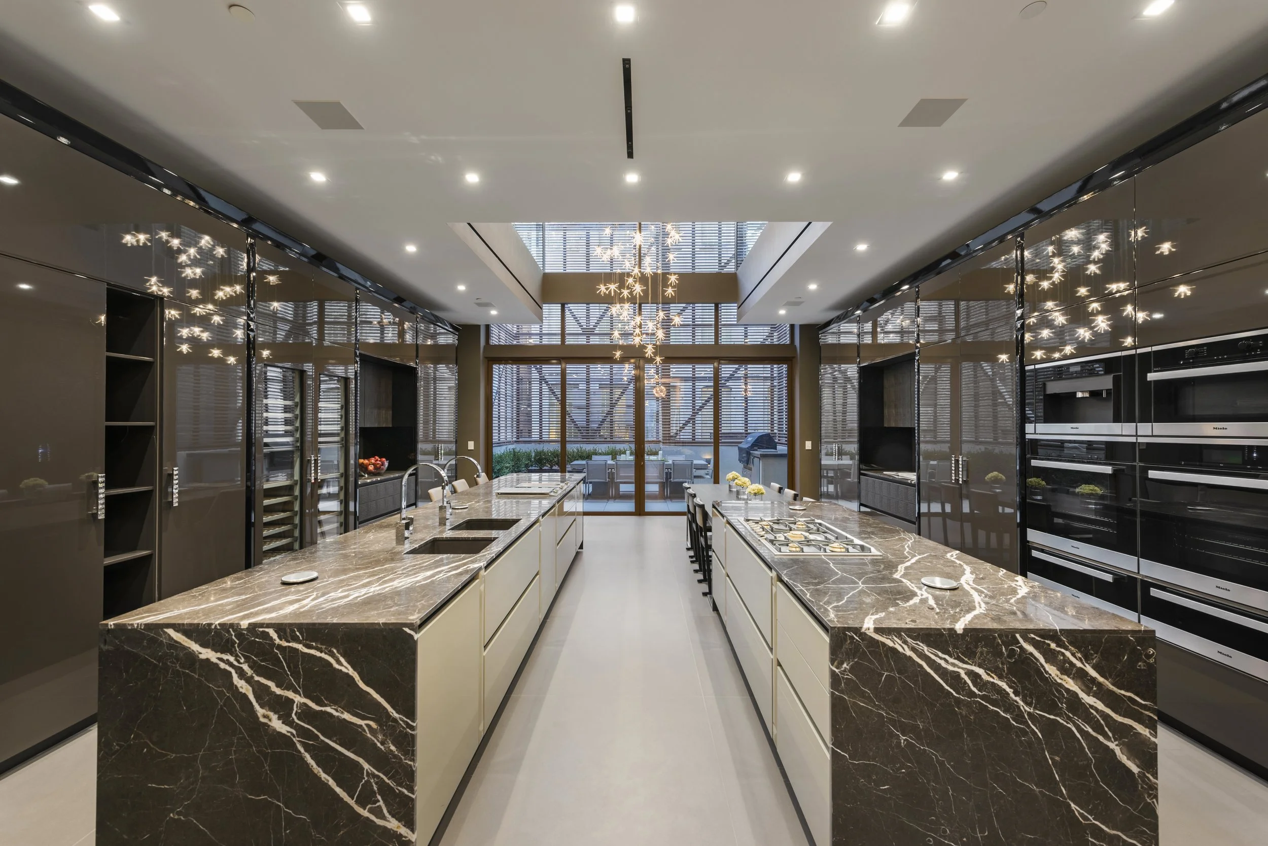 Modern kitchen with marble countertops, multiple ovens, and a large dining area. The room has high ceilings, chandeliers, and large windows with blinds, providing natural light.