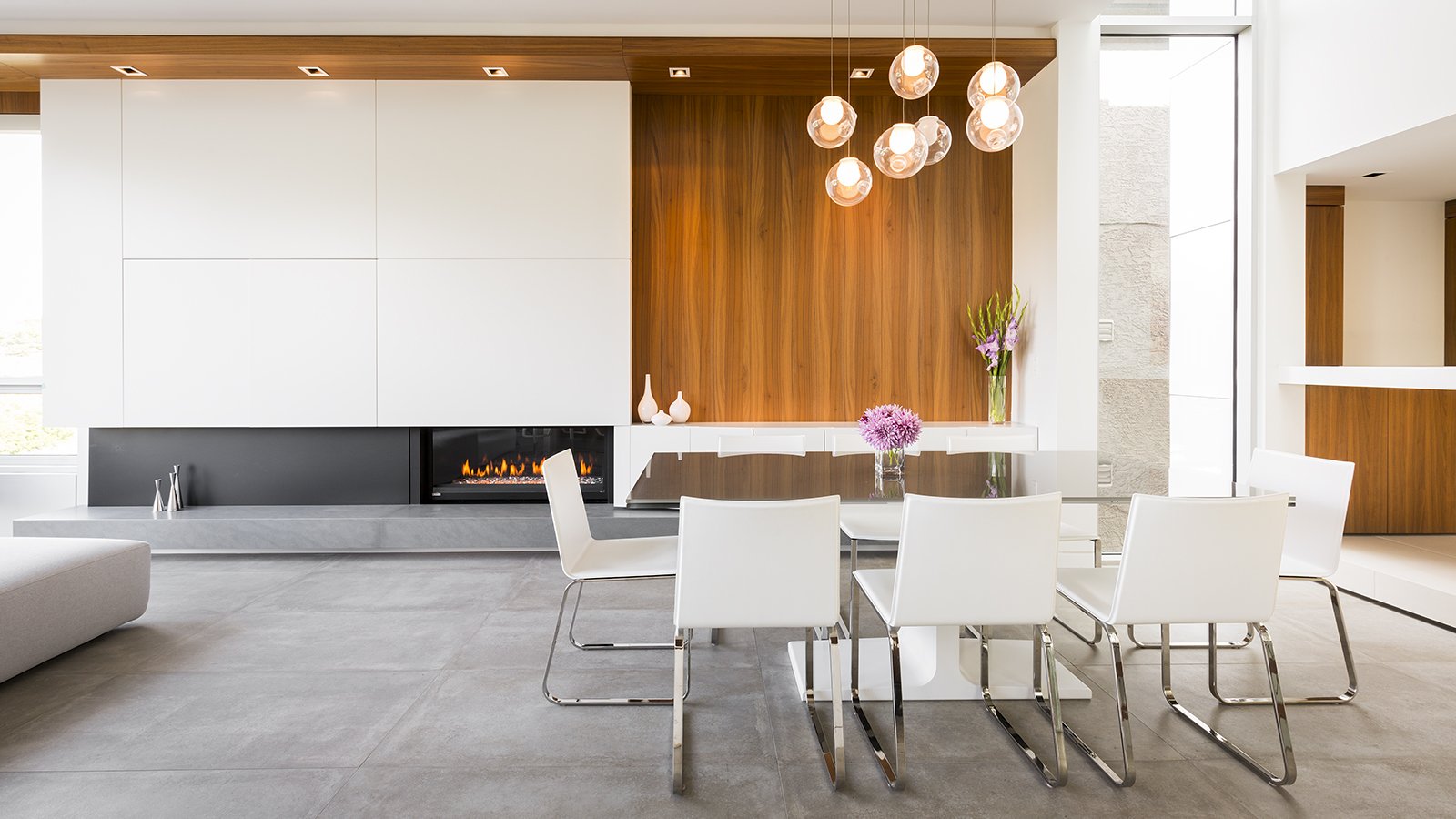 Modern dining room with a glass table, white chairs, a wooden accent wall, and a gas fireplace