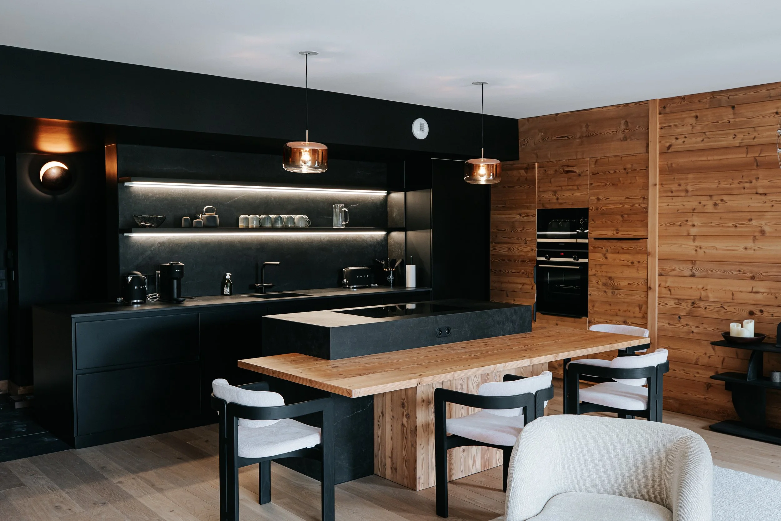Modern kitchen with black cabinets, wooden wall, and a wooden dining table with white and black chairs.