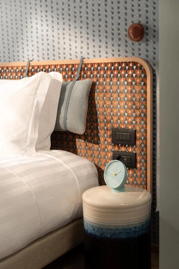 A hotel bedroom scene featuring a bed with white linens, a patterned headboard, a small bedside table with a light blue clock, and a textured wall.
