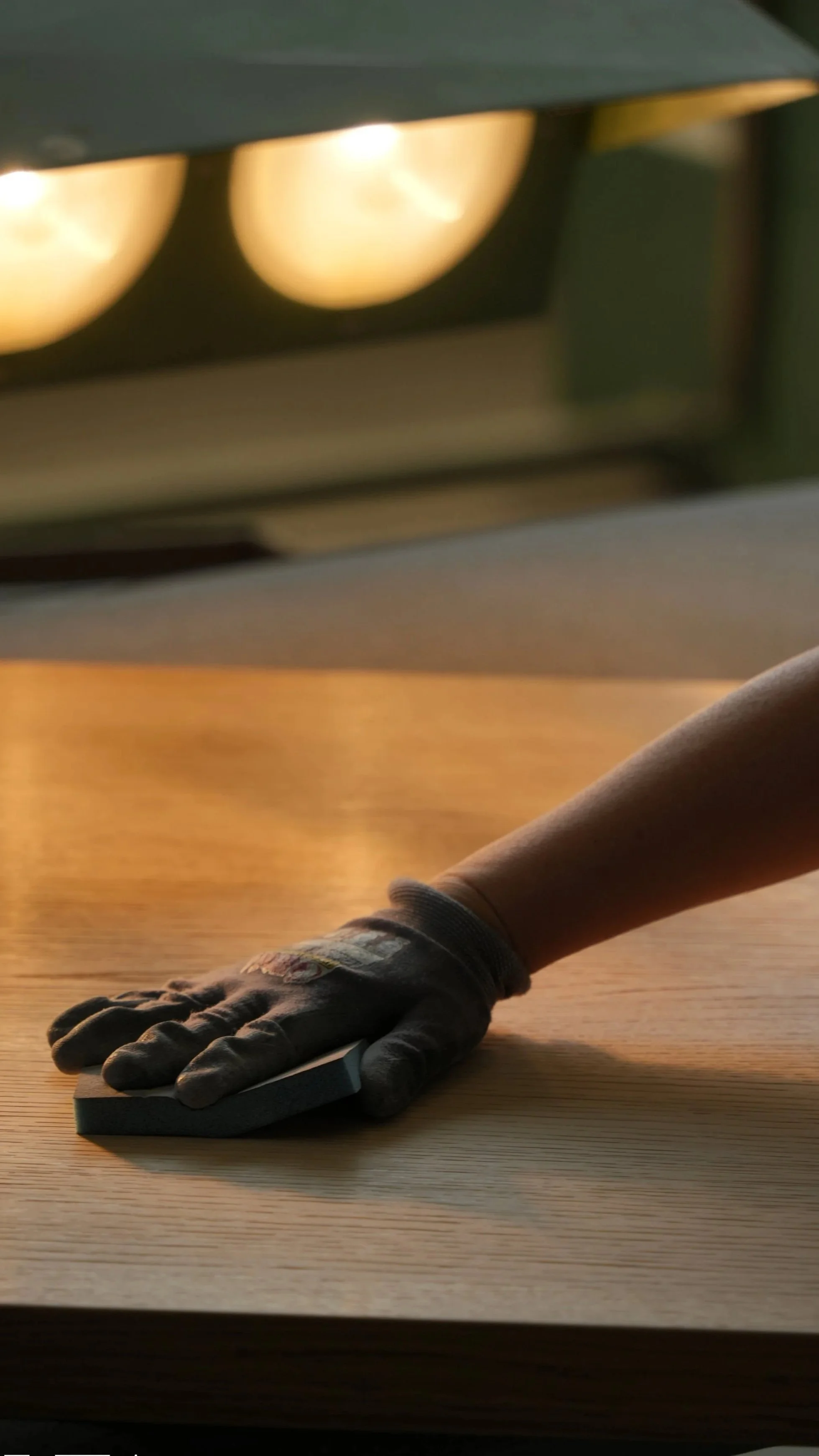 Person wearing black gloves cleaning a wooden surface with a cloth in a cabinet factory setting.