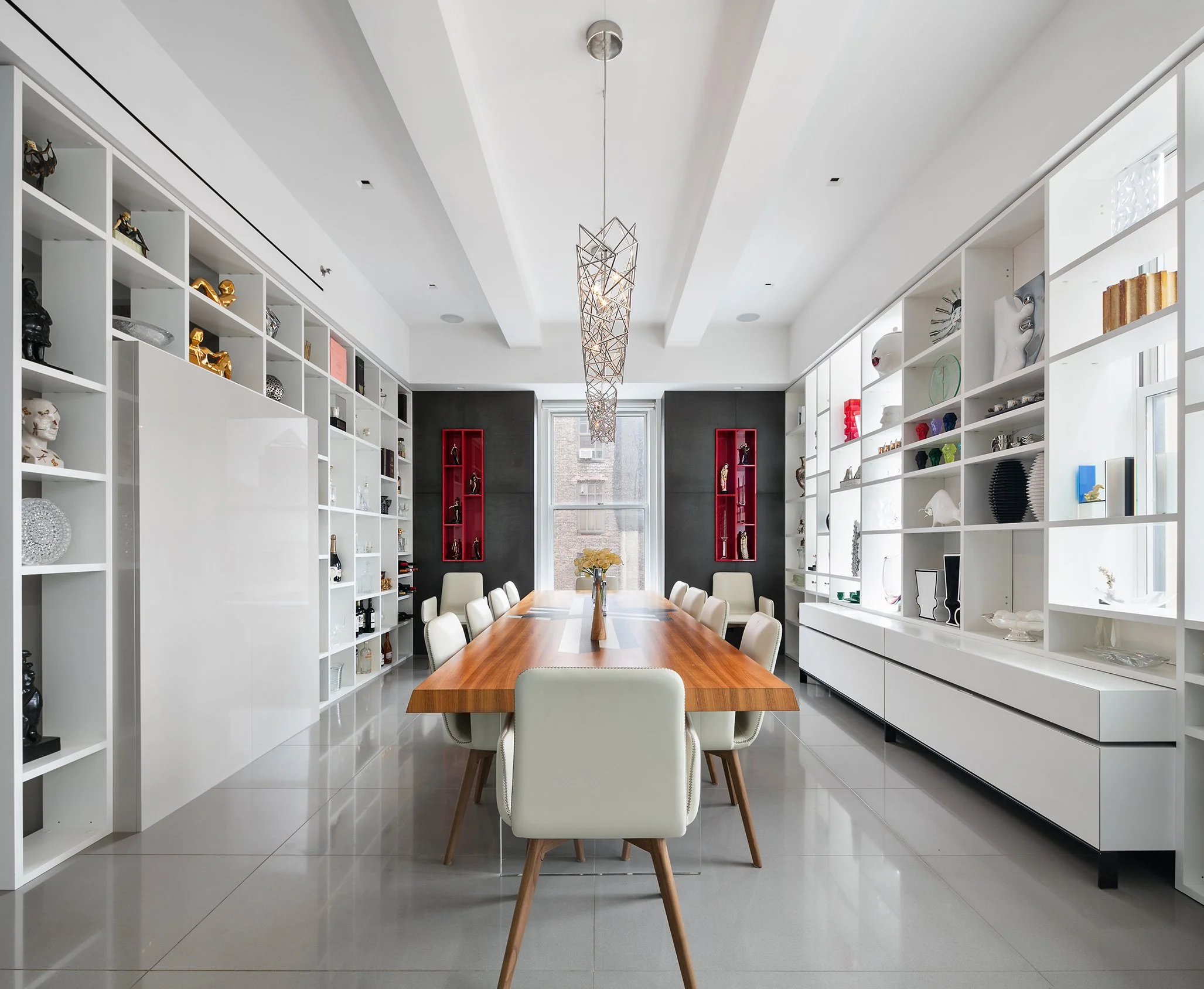 Modern dining room with white built-in bookshelves displaying decorative items, a wooden dining table with white chairs, and large windows letting in natural light.