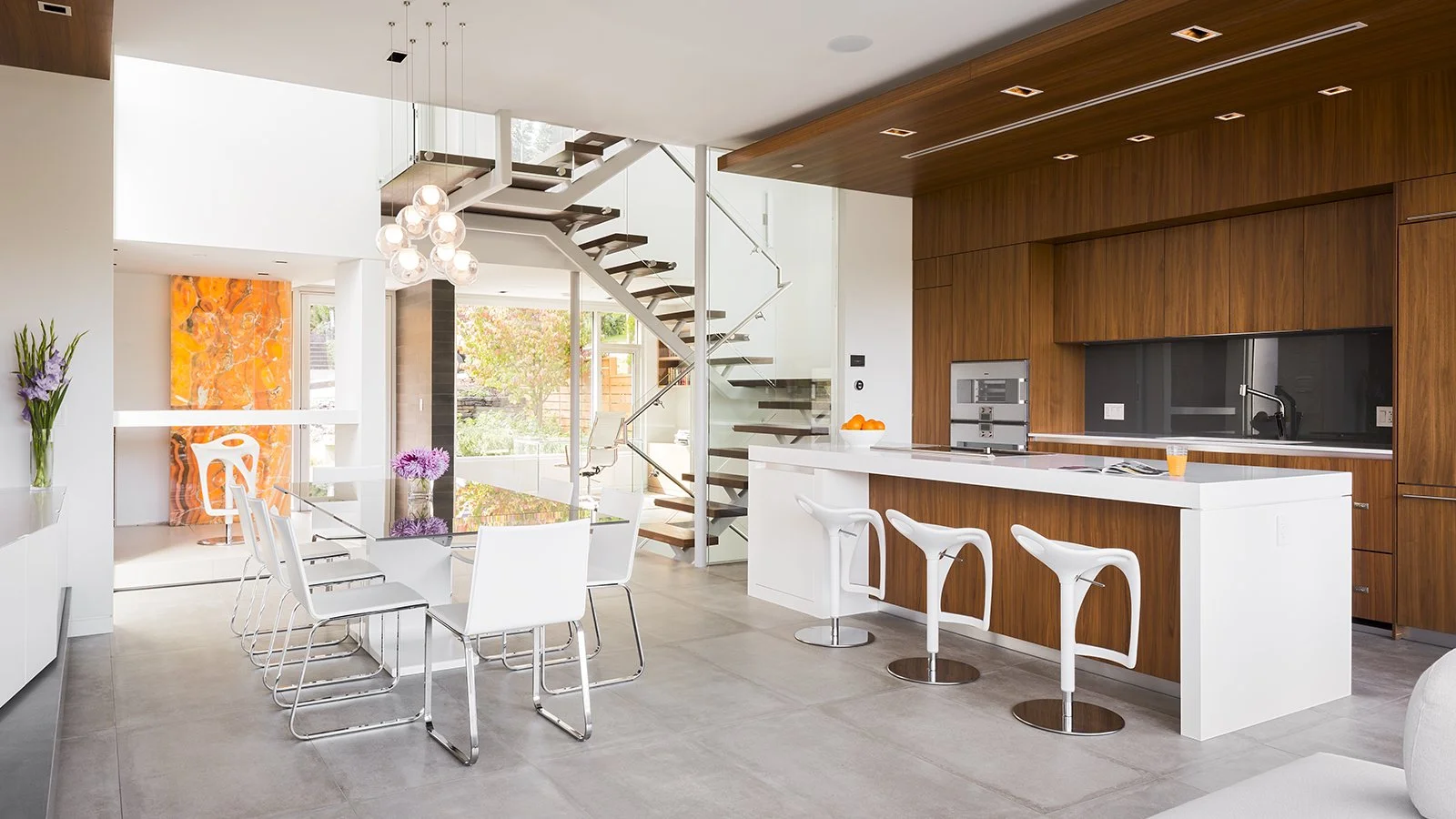 Modern, luxury open-concept kitchen and dining area with white and wood accents, a glass dining table with white chairs, a kitchen island with three white barstools, a staircase with glass railing, and large windows letting in natural light.