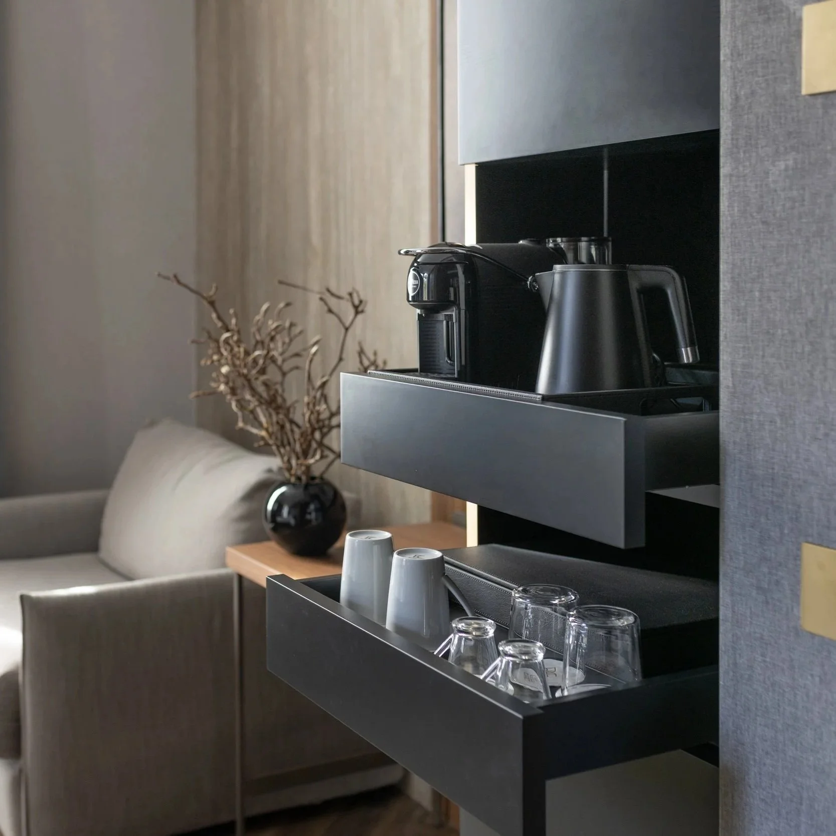 A view of a modern kitchen with a coffee machine, a black kettle, and a tray with four glasses and two cups on a black shelf, with a beige sofa, armchair, wooden side table, and a decorative branch in a black vase in the background.