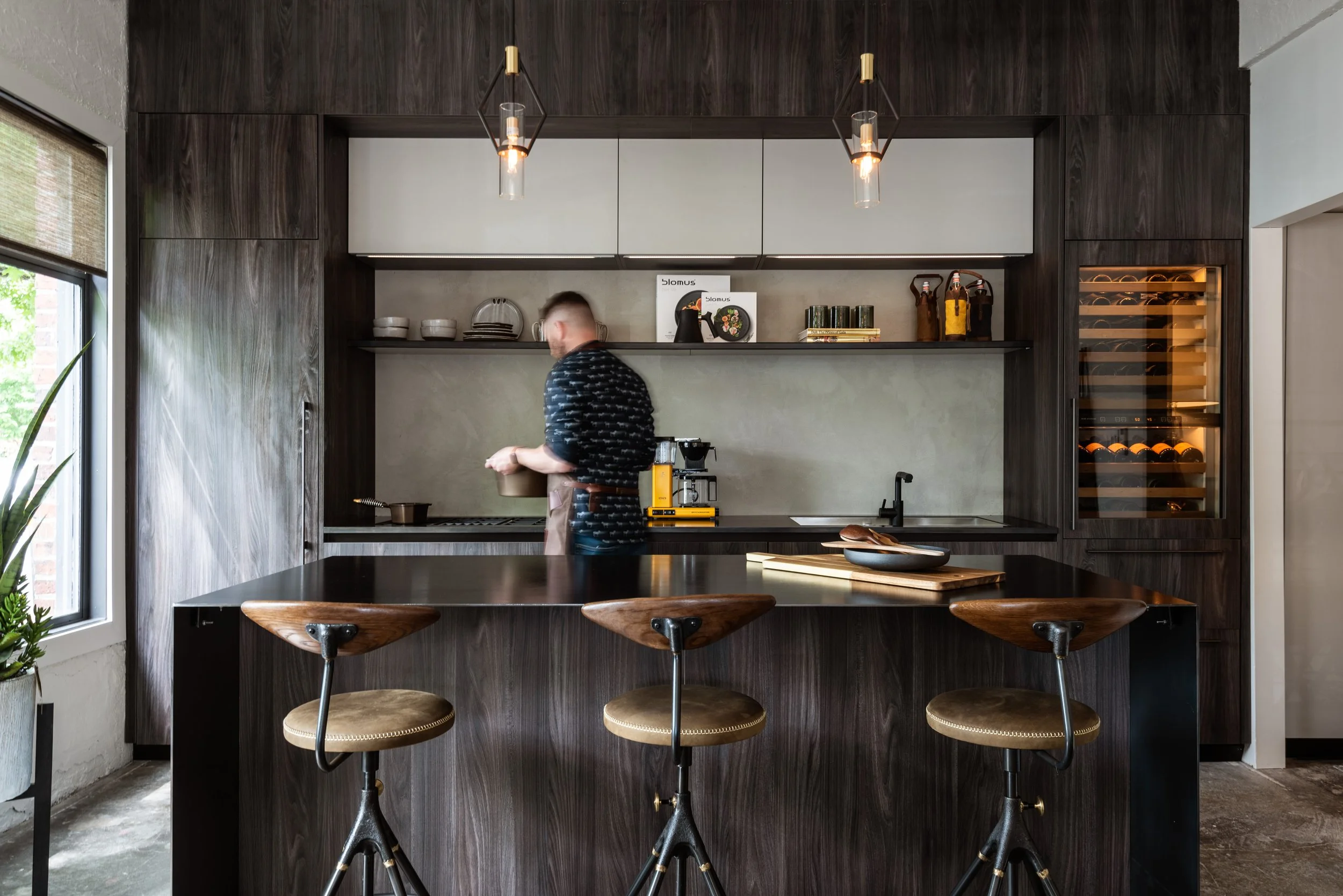 Modern kitchen with dark wood cabinetry, a concrete backsplash, and a large window on the left. A man is standing at the counter, preparing coffee. There are three brown barstools with metal bases at the kitchen island, which has a chocolate bowl and a cutting board on it. Pendant lights hang above the counter, and a wine cooler is built into the cabinetry on the right.