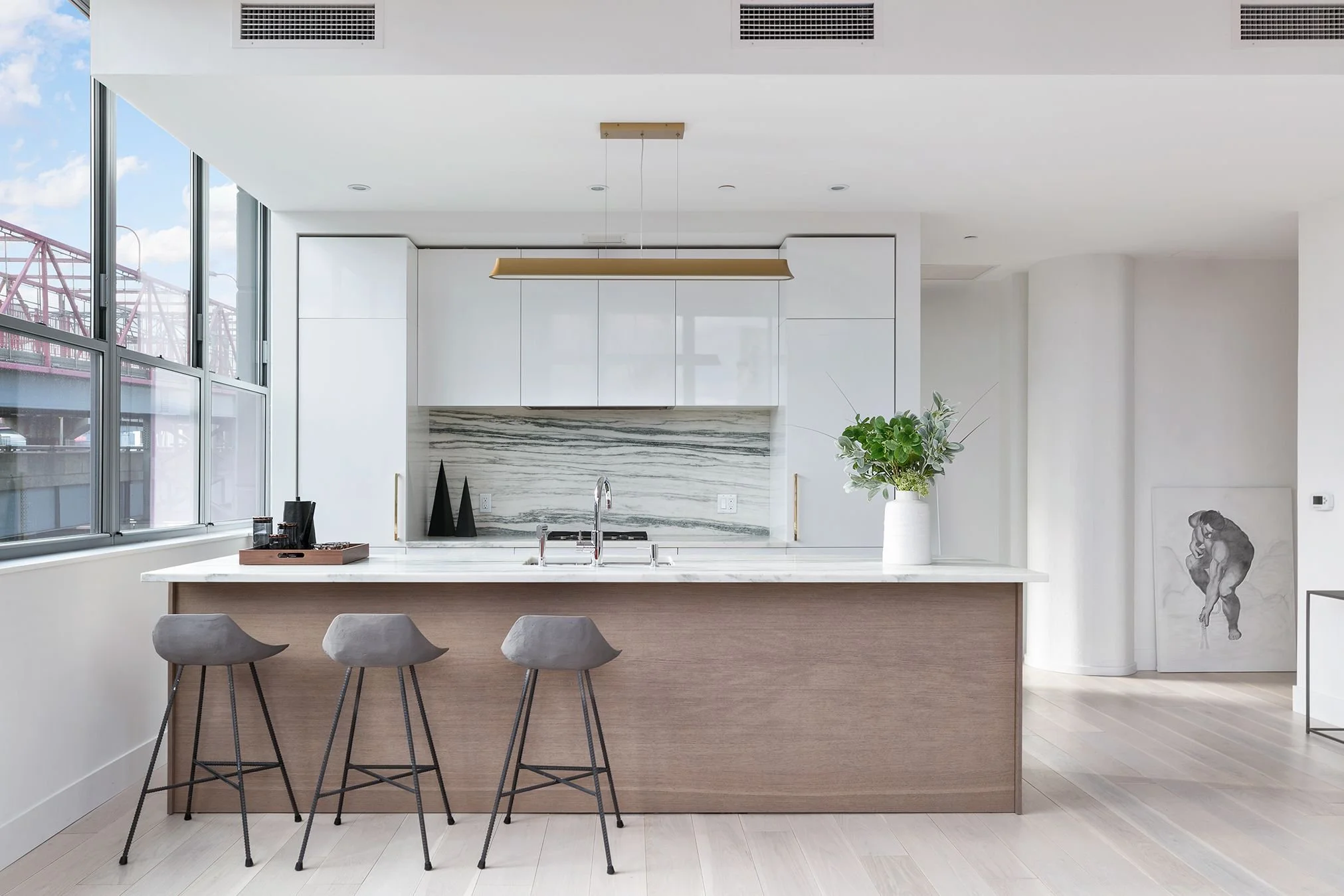 Modern kitchen with a marble island, white cabinets, and large windows showing a bridge outside. Decor includes black vases, a green potted plant, and a dog illustration on the wall.