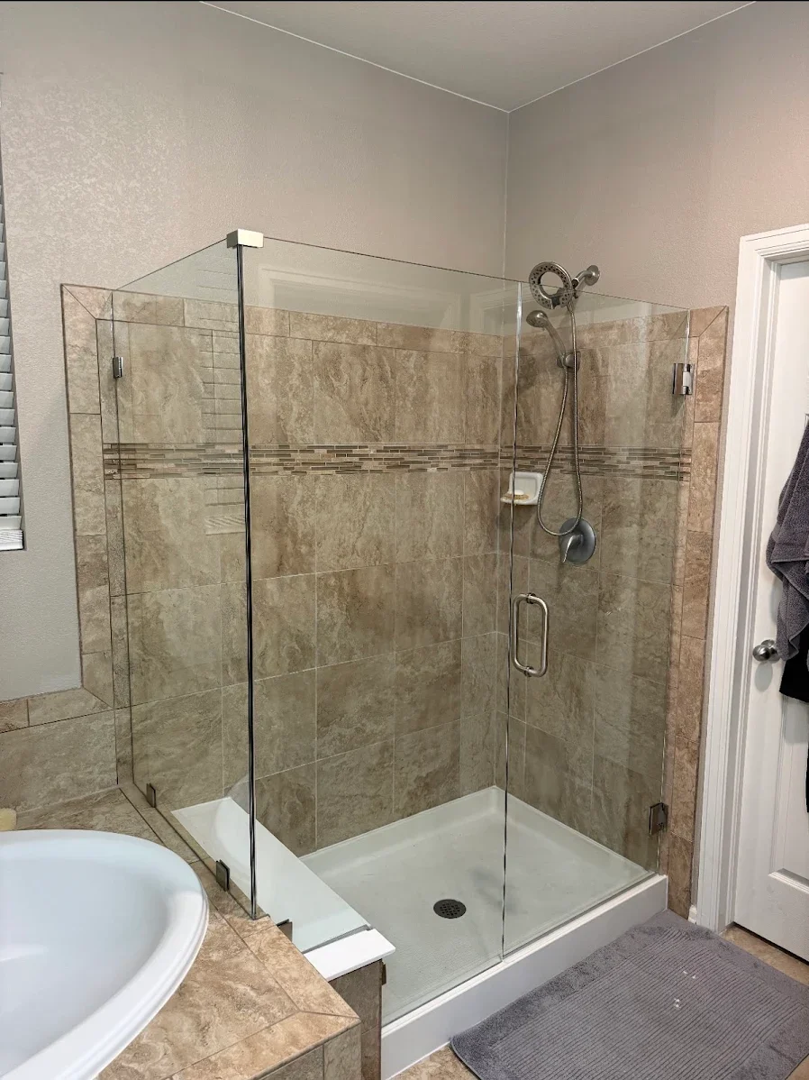 Bathroom shower with clear glass doors, beige tiled walls, and a white shower base. There is a handheld showerhead and small soap dish inside. Part of a white sink and a gray towel hanging on the door are visible.