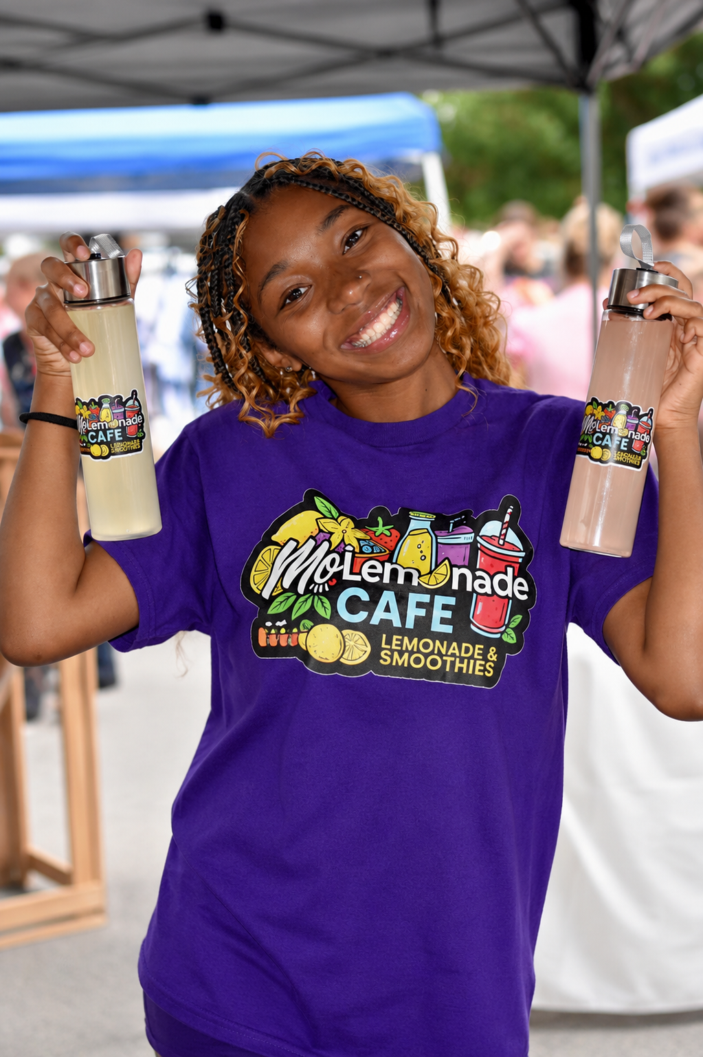 A smiling woman in a purple T-shirt with a Lemonade Cafe logo, holding two bottles of lemonade at an outdoor event.