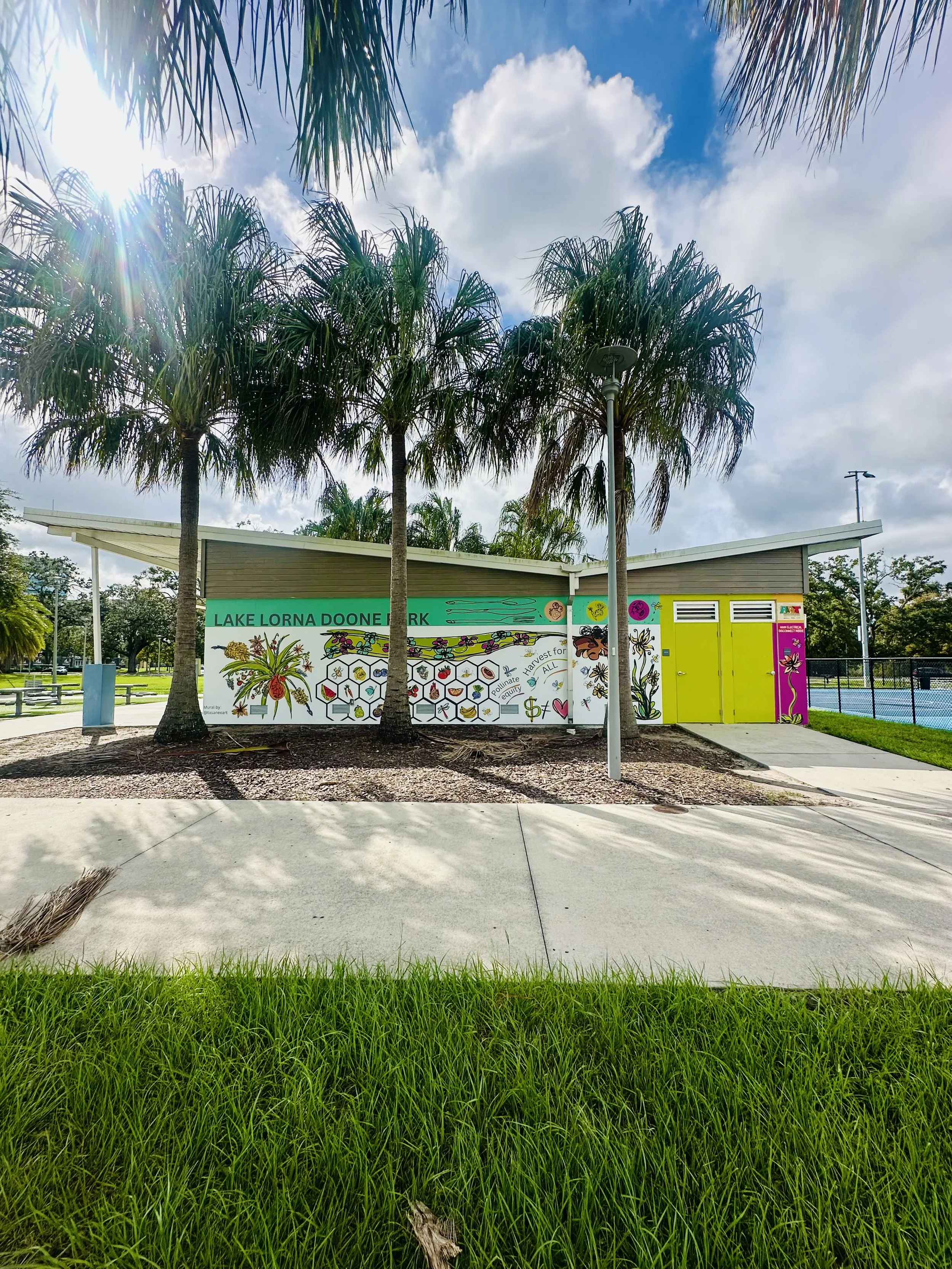 Colorful informational sign at Lake Lorna Doone Park with paintings of plants and bees, surrounded by green grass and tall palm trees under a partly cloudy sky.