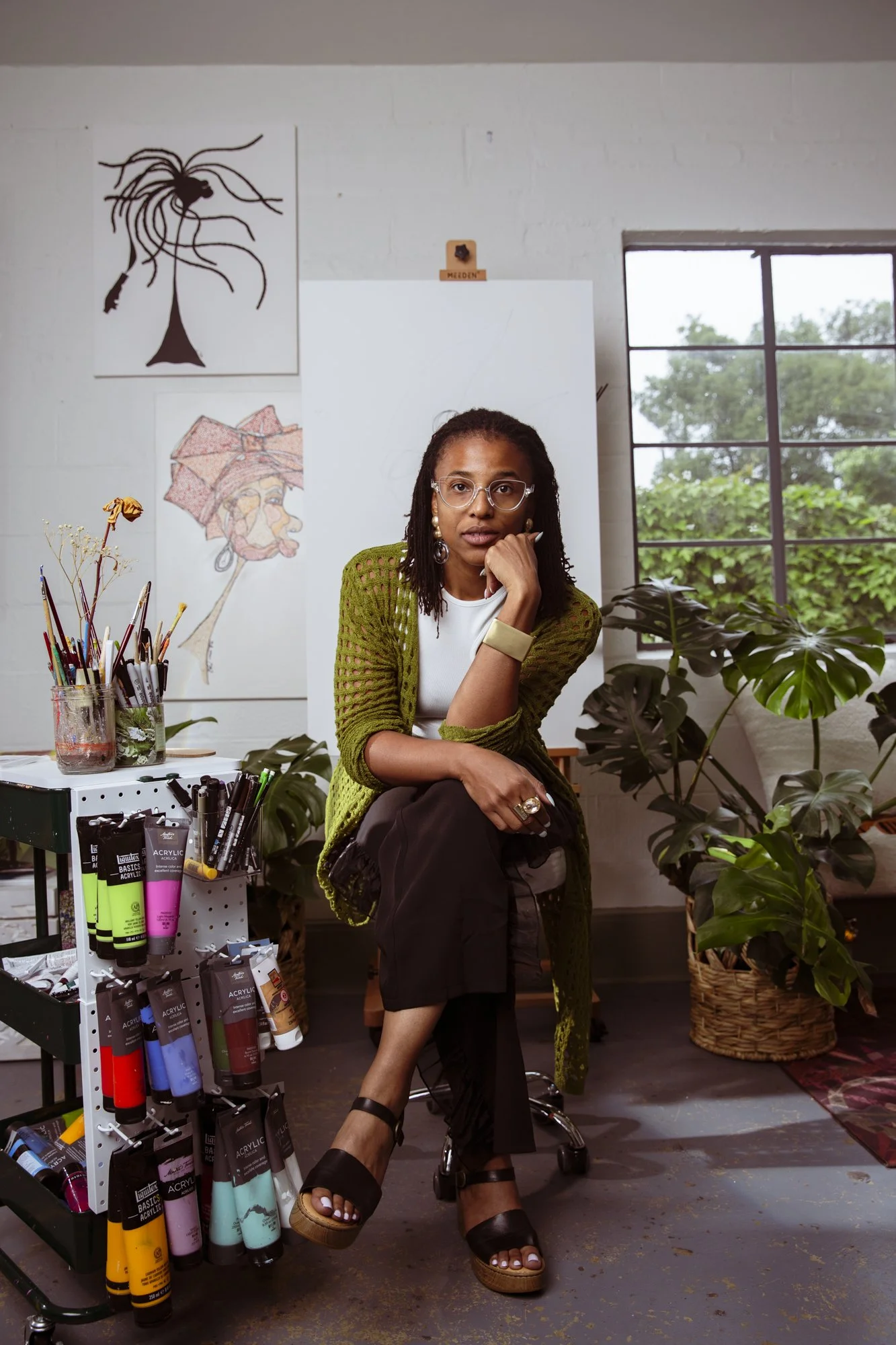 A woman with glasses and dreadlocks sitting in an art studio surrounded by plants and art supplies, including paints and markers, with paintings on the wall and a window showing greenery outside.