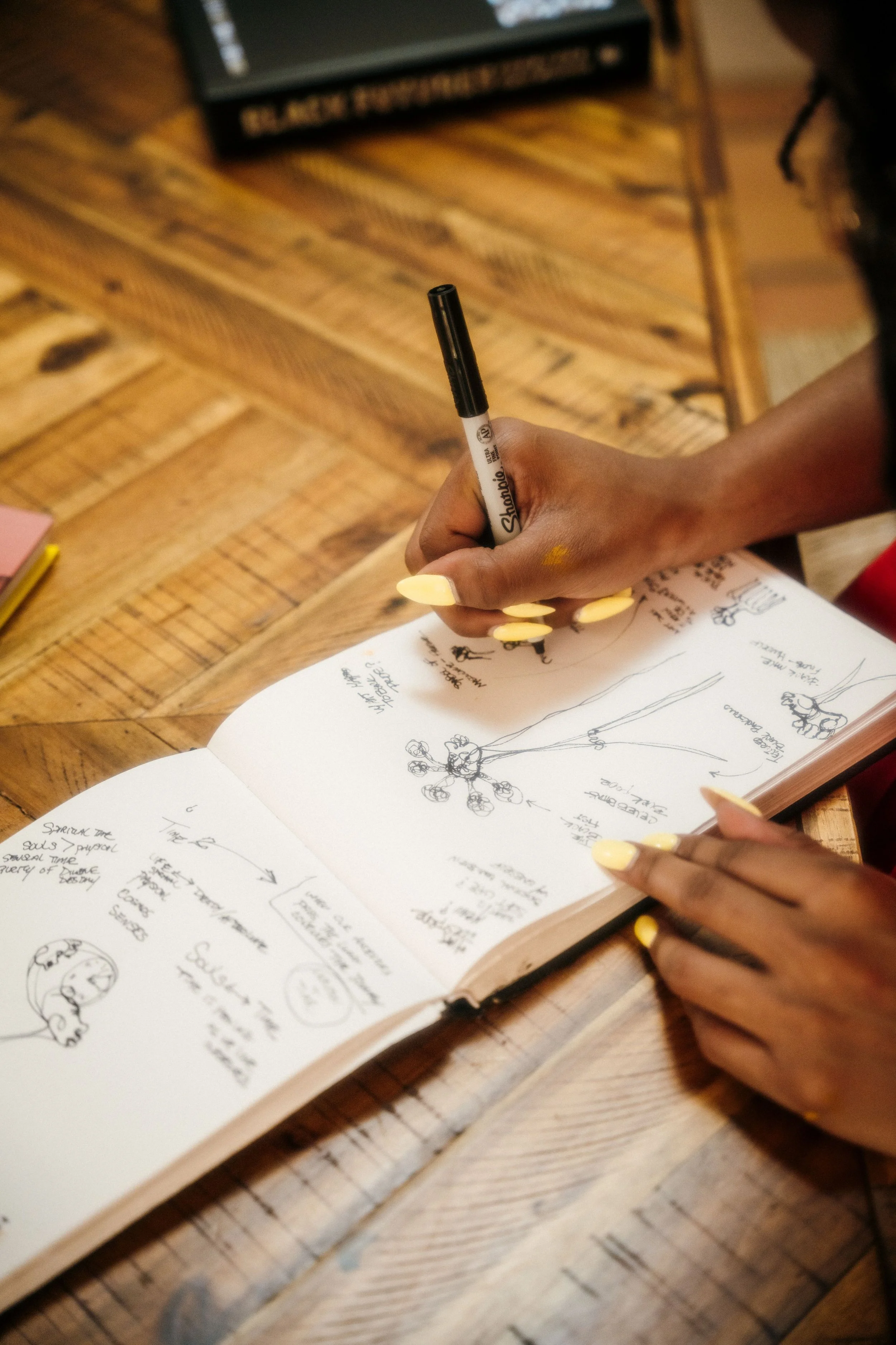 A person with yellow-painted nails is drawing diagrams and notes in a sketchbook with a black marker, on a wooden table.