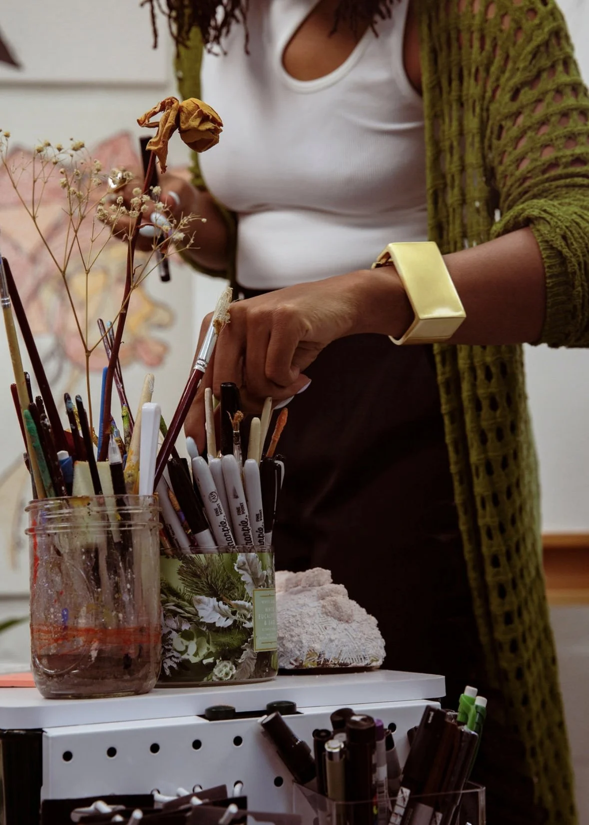Person in a white tank top and green cardigan selecting pens or markers from a container, with various art supplies and dried flowers on a table.