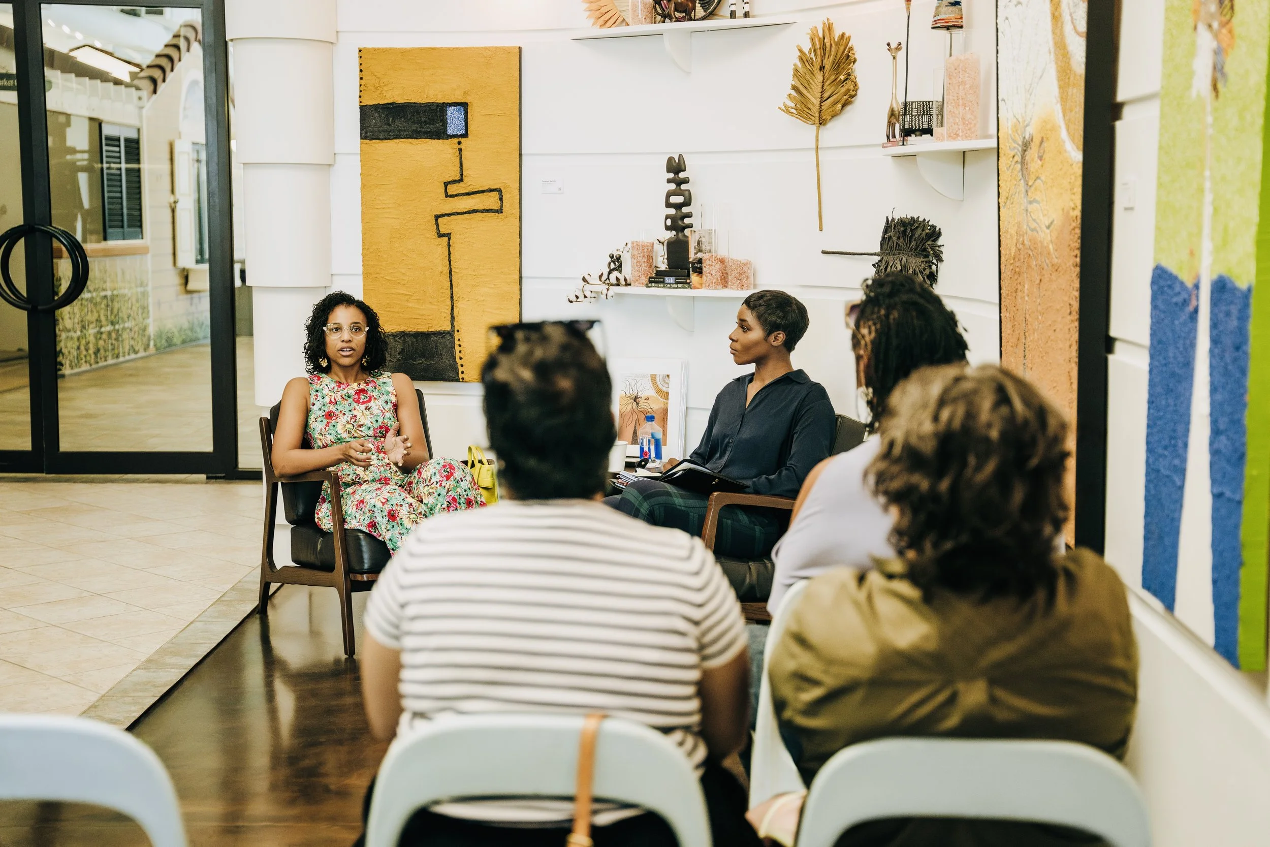 A group of people seated in a room, engaged in a discussion or meeting. The room features colorful abstract artwork on the wall and a white shelf with decorative items. The image is viewed through a glass door.