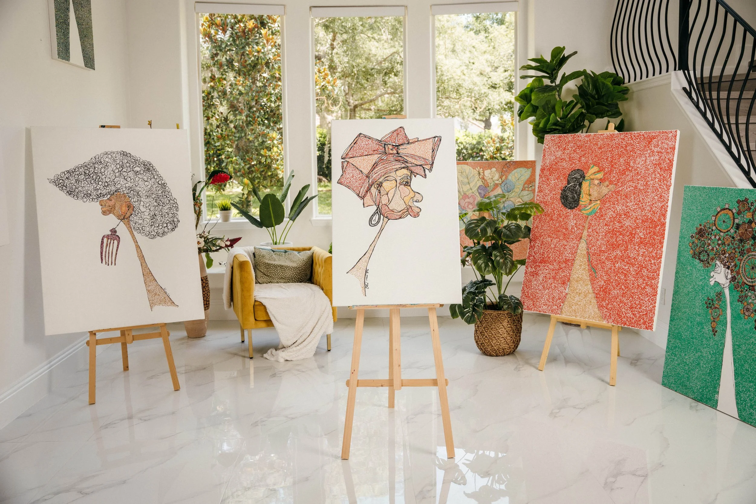 An art gallery displaying colorful paintings on easels in a bright room with large windows and a view of greenery outside.