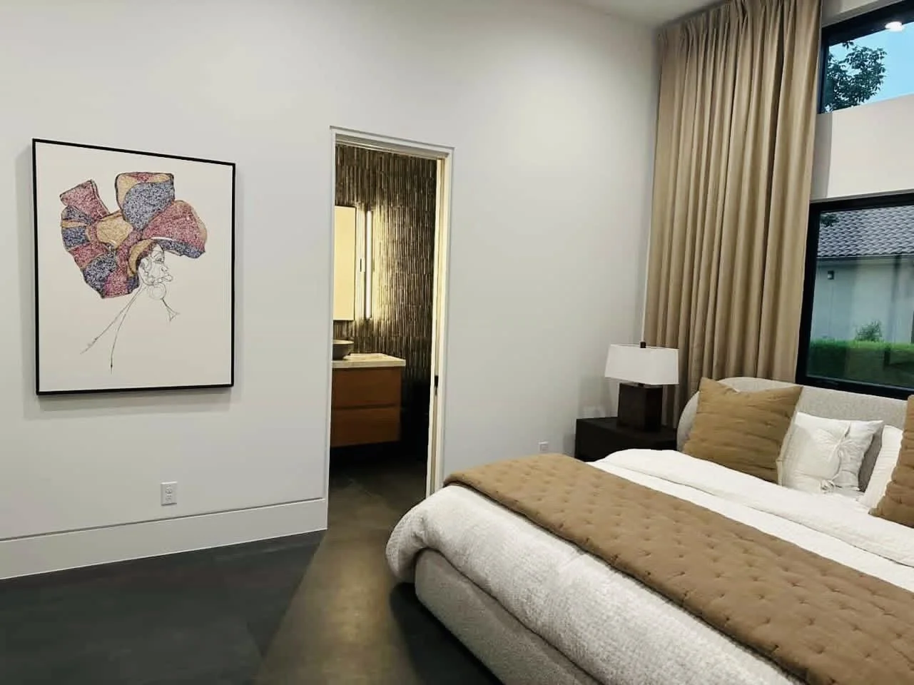 Modern bedroom with a large bed, beige and white bedding, and pillows. A bedside table with a lamp, window with curtains, and artwork on the wall.