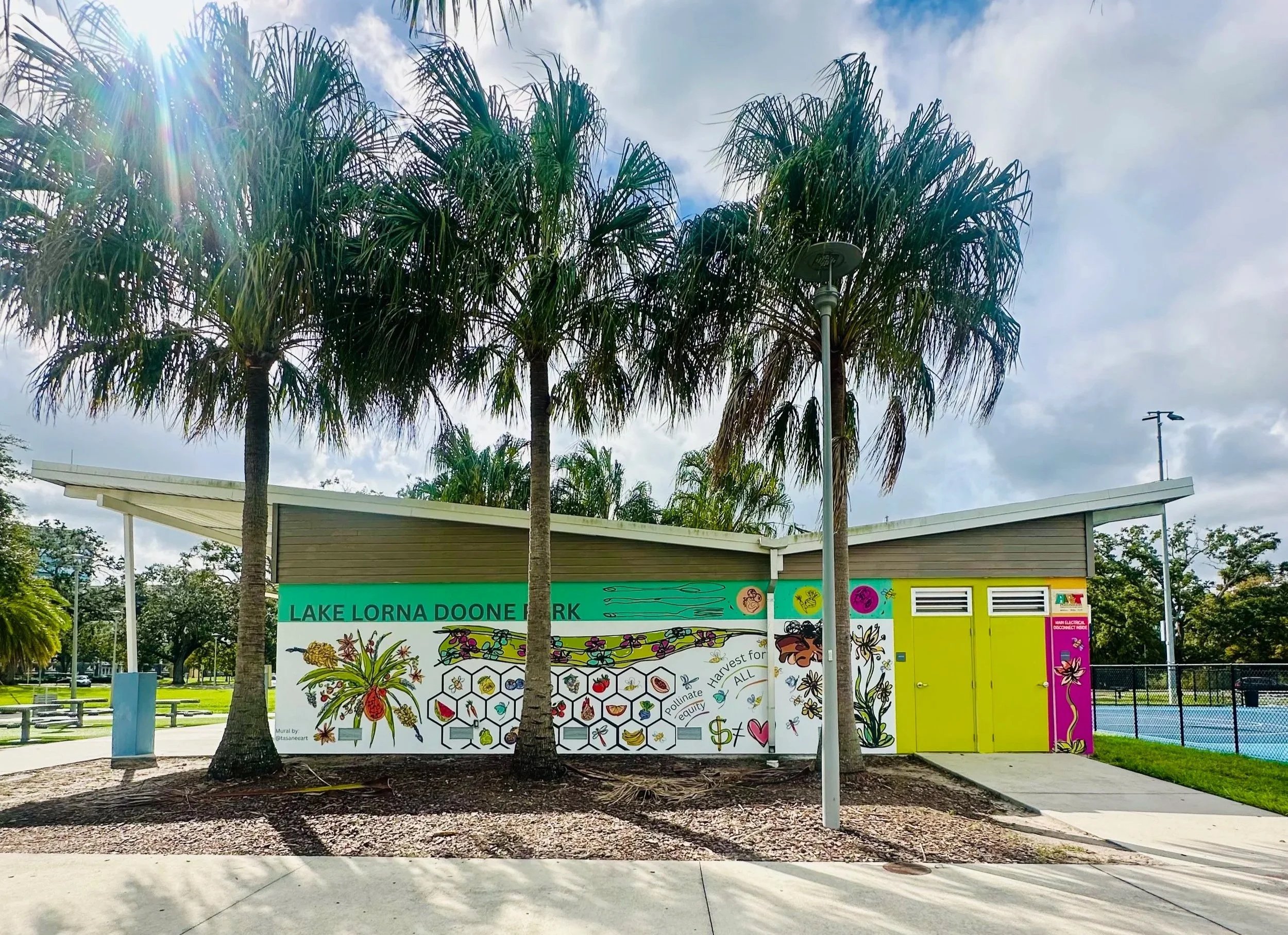 A colorful educational mural about pollinators and sustainable harvesting at Lake Lorna Doone Park, with palm trees, a bright sky, a park building, and a tennis court in the background.