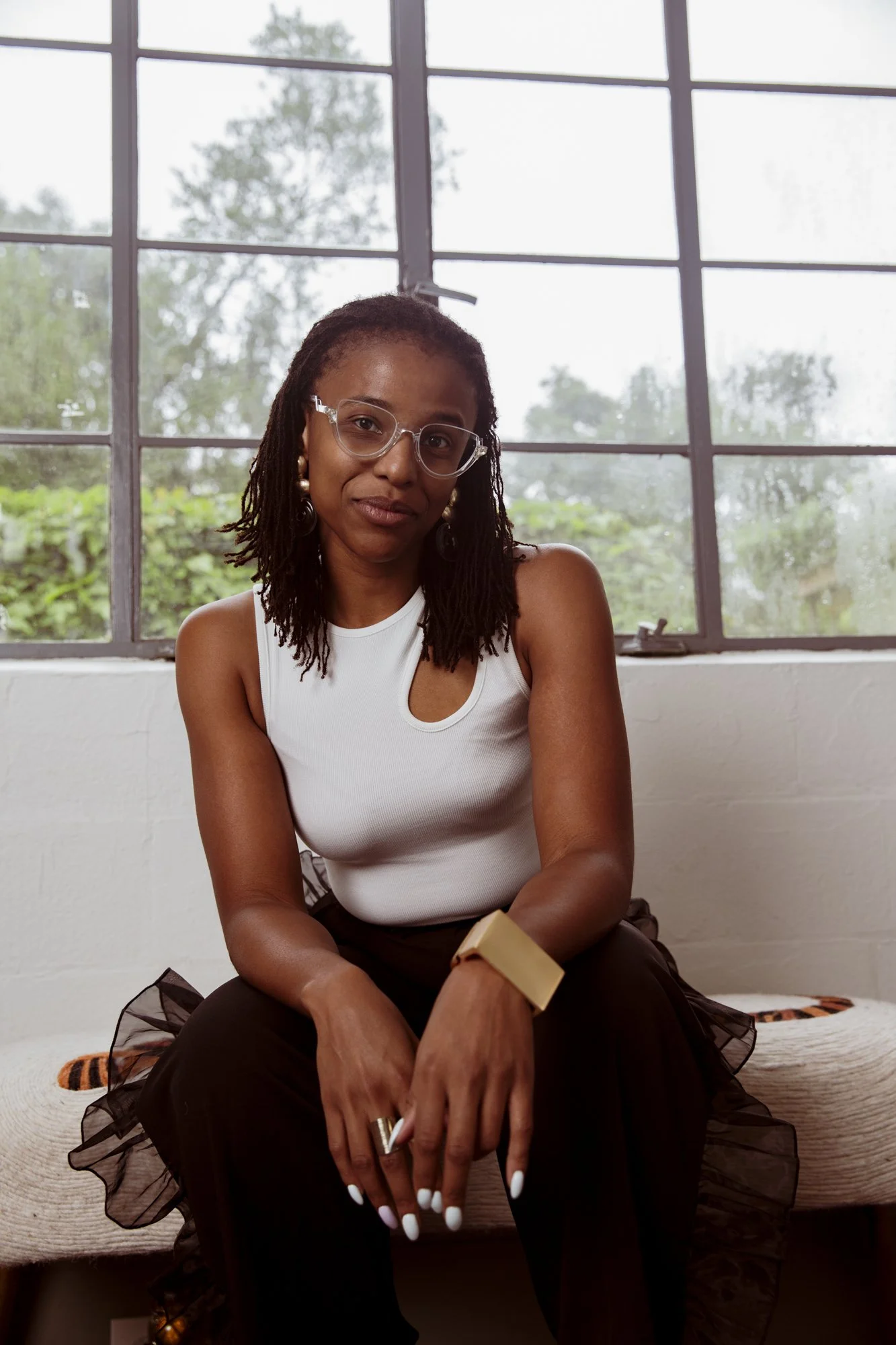 A woman with glasses and earrings sitting by a large window, wearing a white sleeveless top, black skirt with sheer ruffle detail, holding a notebook or book, with greenery outside the window.