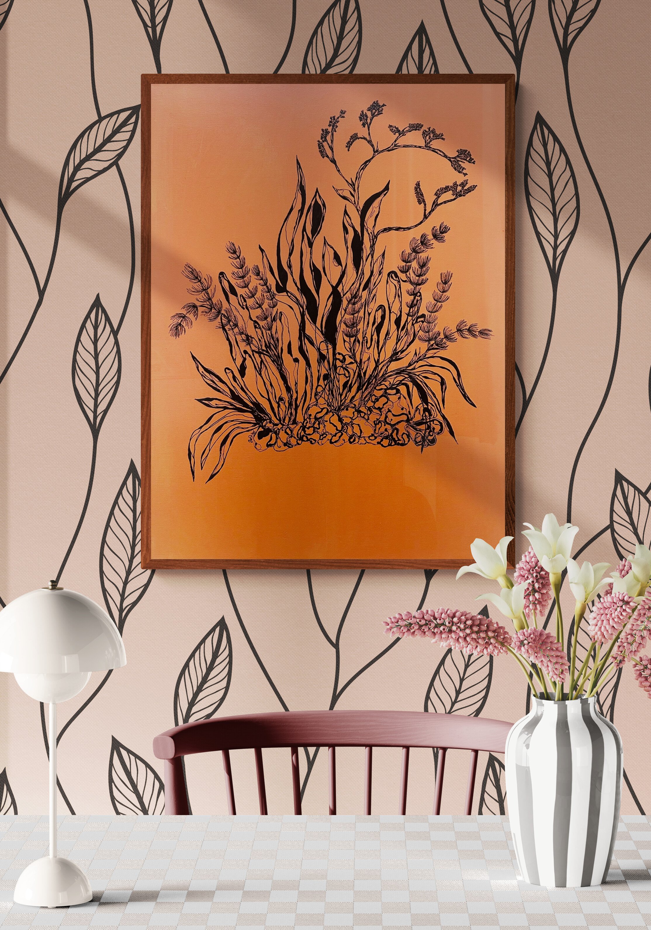 Interior of a room with a pink wall featuring a black leaf pattern, a framed artwork of a black botanical line drawing on an orange background, a white table lamp, a pink chair, and a vase of pink and white flowers on a checkered table surface.