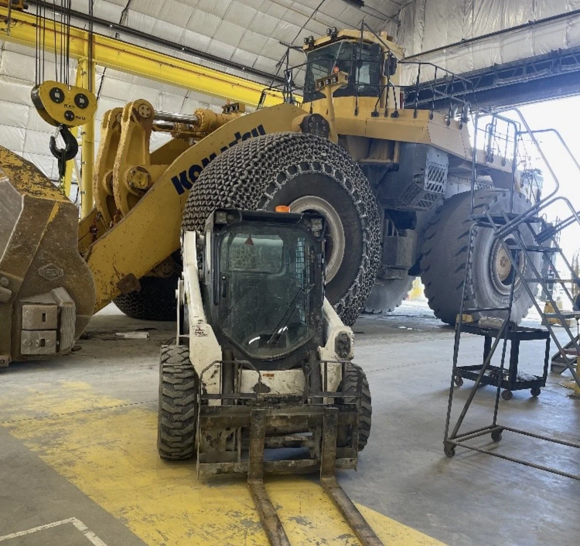 A large Komatsu construction vehicle inside a warehouse with high ceilings, accompanied by a smaller forklift in the foreground and a mobile platform with steps on the right.